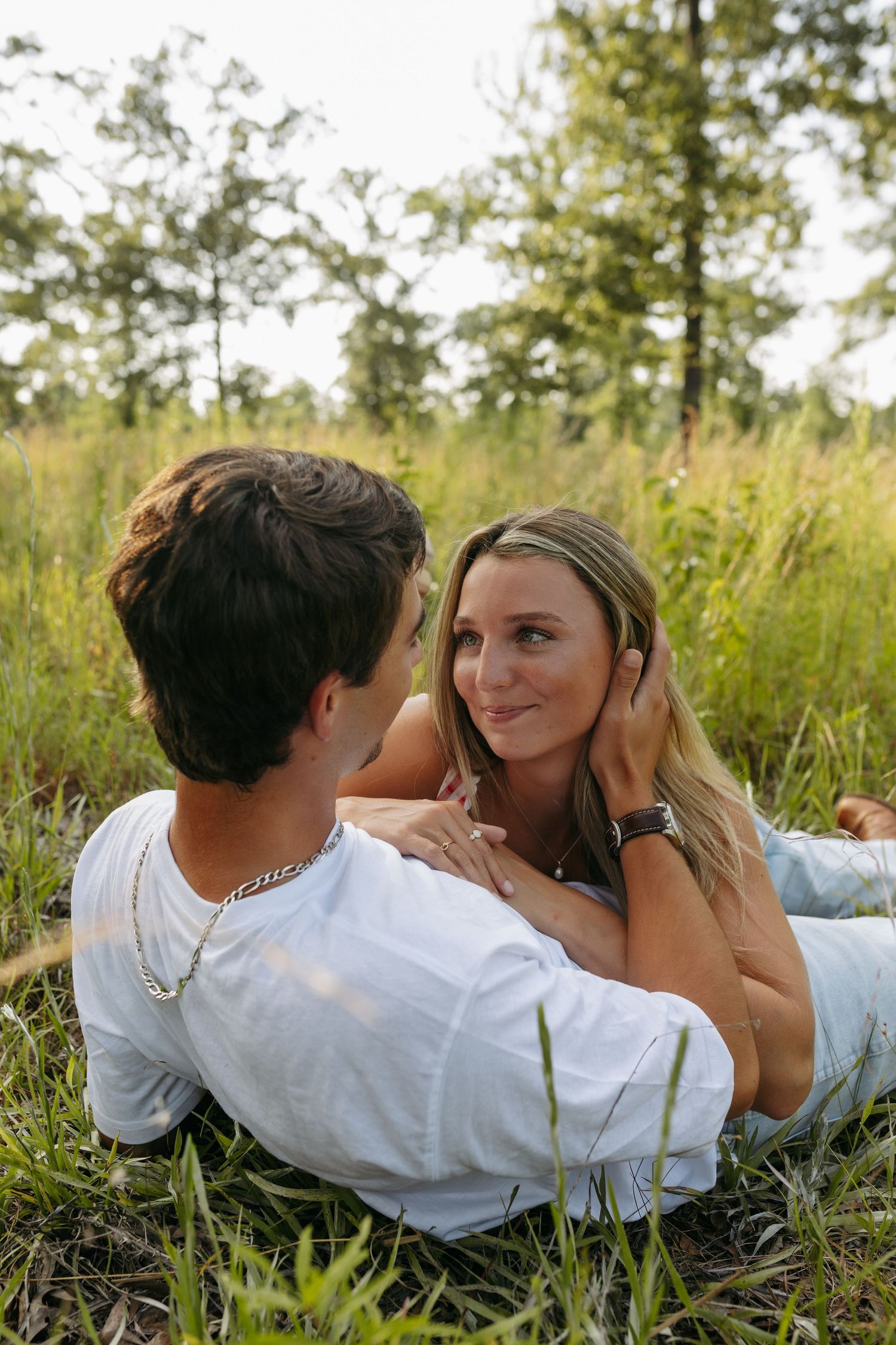 A young couple lying in green grass in a forest, gazing at each other romantically during golden hour.
