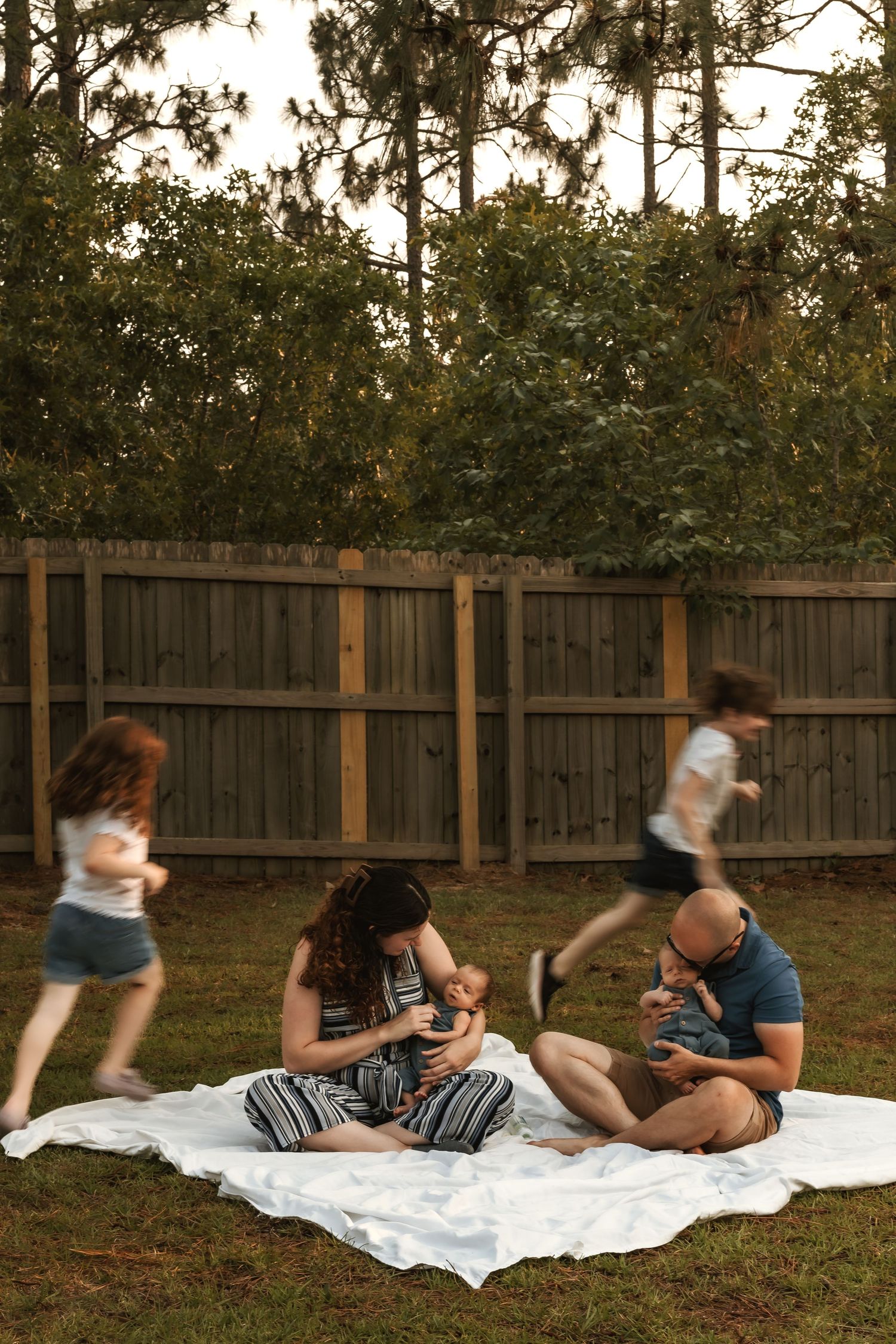 Family relaxing together on white blanket in fenced backyard at sunset.