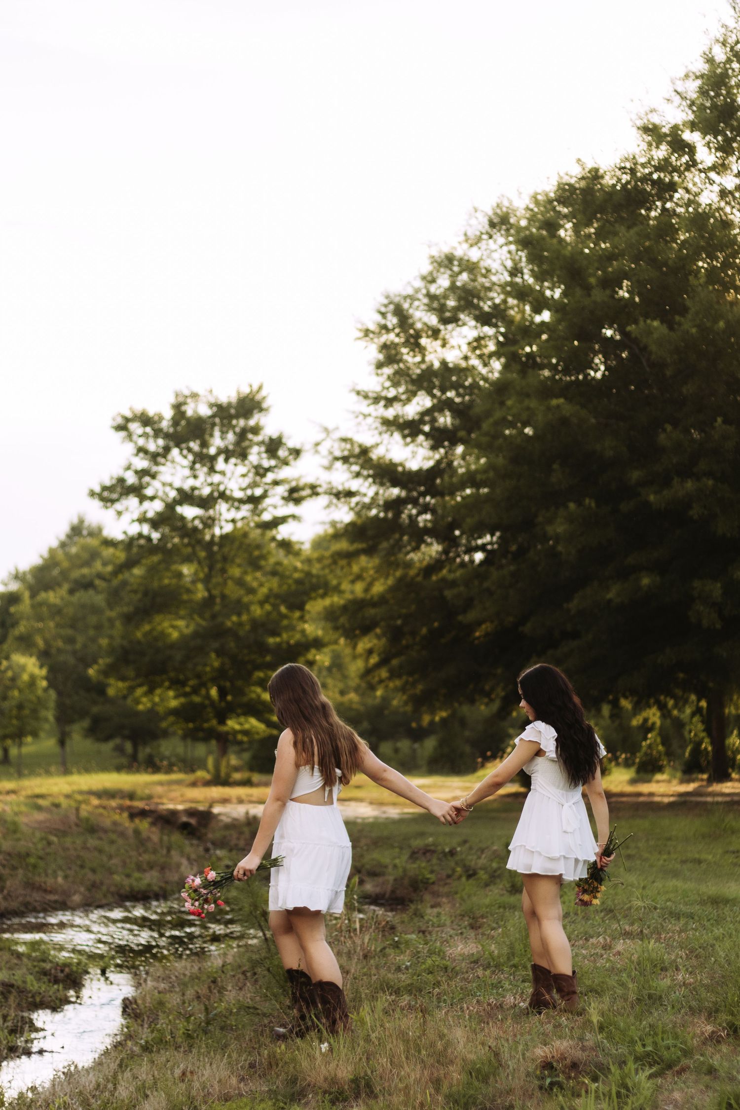 Two people in white dresses and boots hold hands while walking together near a stream at sunset in a grassy field.
