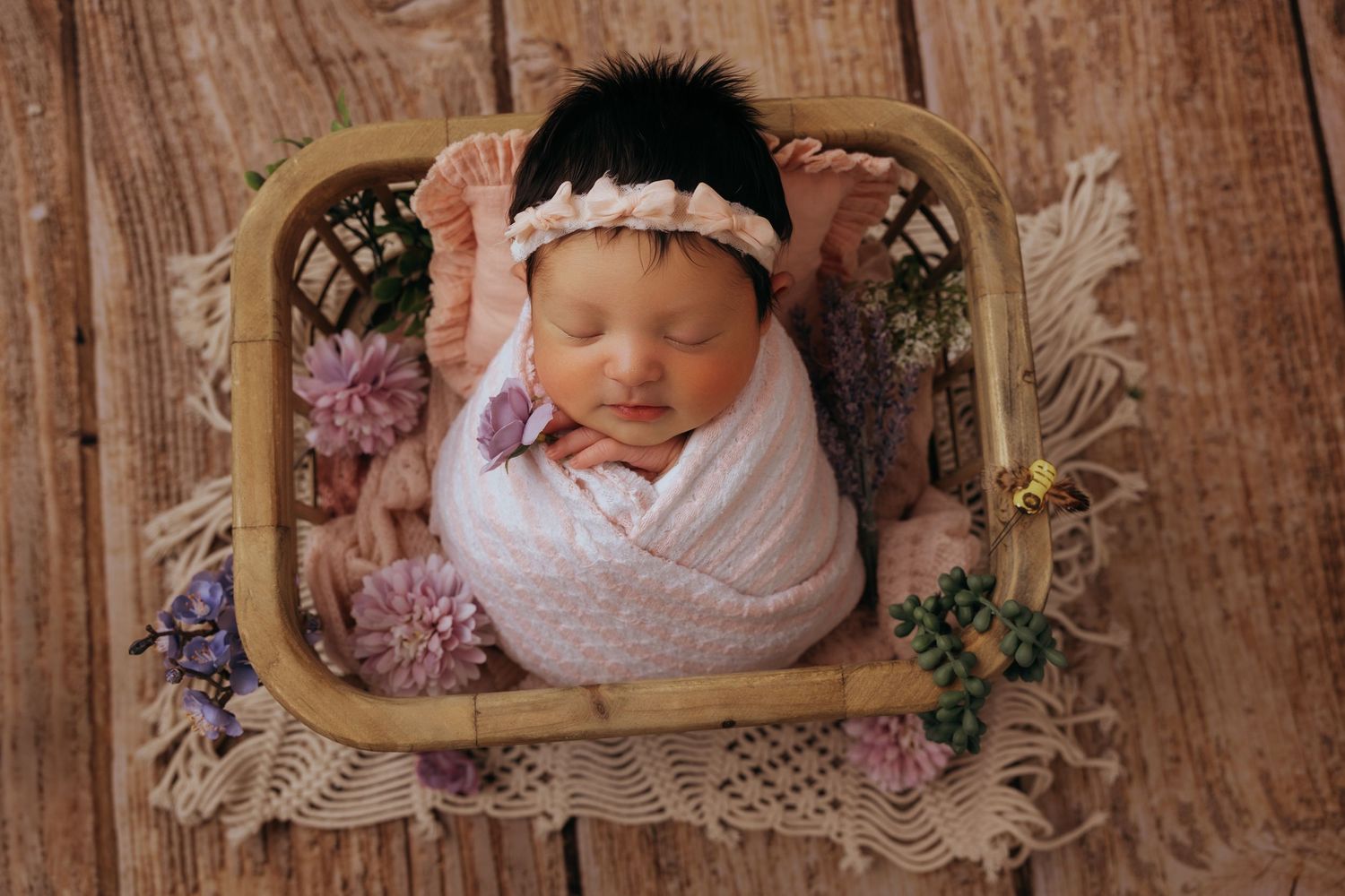 Newborn baby wrapped in pink swaddle sleeps peacefully in vintage basket with dried flowers on wooden backdrop.