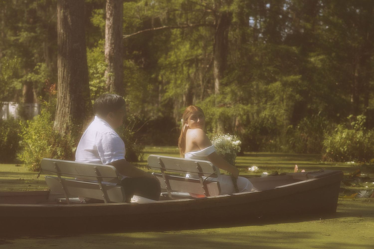 Couple sitting in a wooden canoe on a serene lake surrounded by lush green trees, woman holding a bouquet.