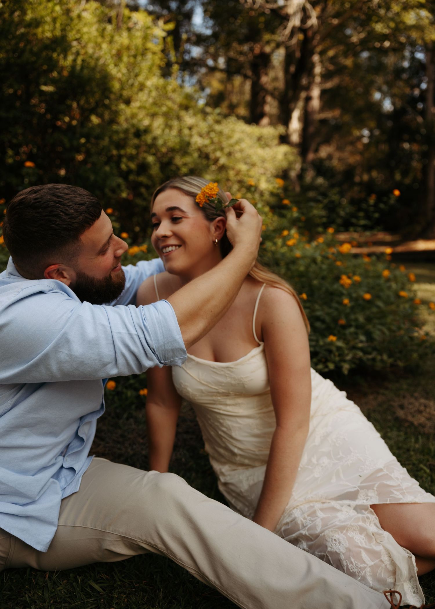 A couple sits in a lush garden as a man gently places an orange flower in a smiling woman's hair.