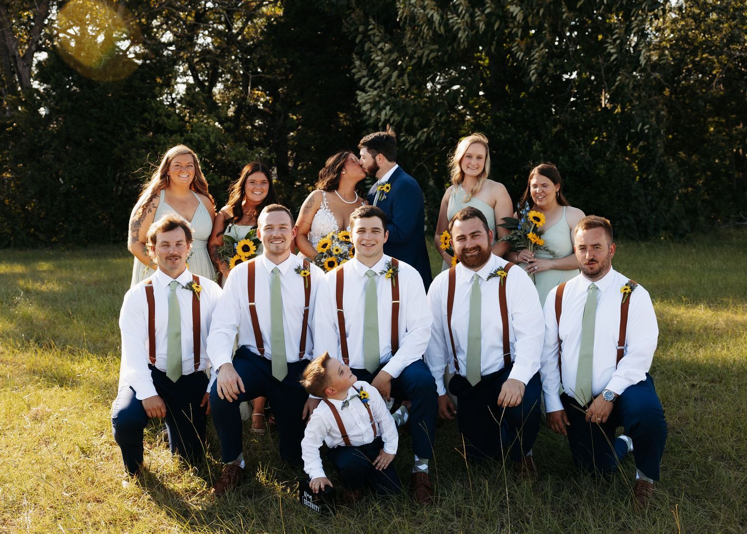 Wedding party poses outdoors, groomsmen in white shirts with suspenders, bridesmaids in sage green, couple kissing behind.