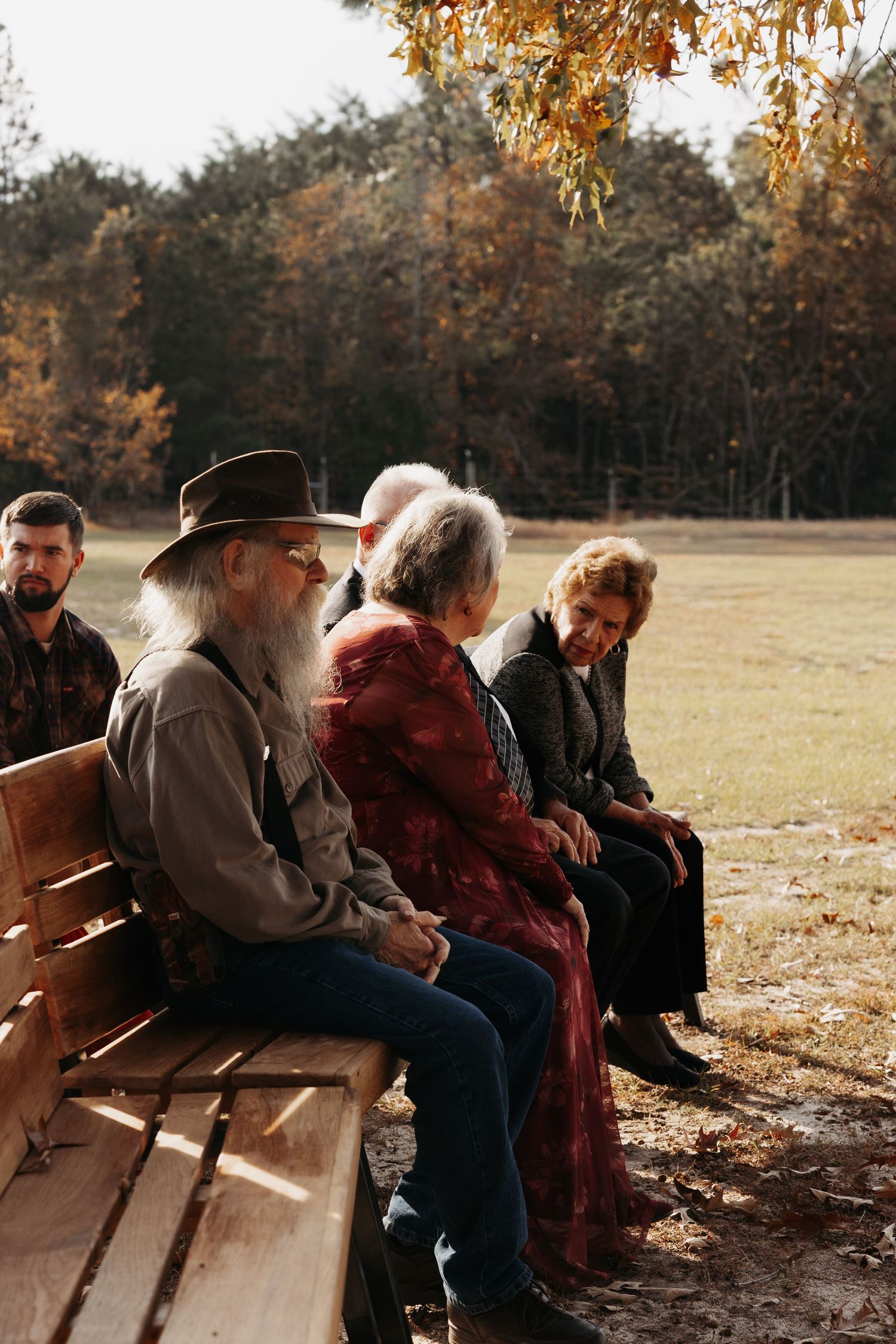 Wedding guests seated on wooden benches outdoors during an autumn ceremony in a rustic park setting.