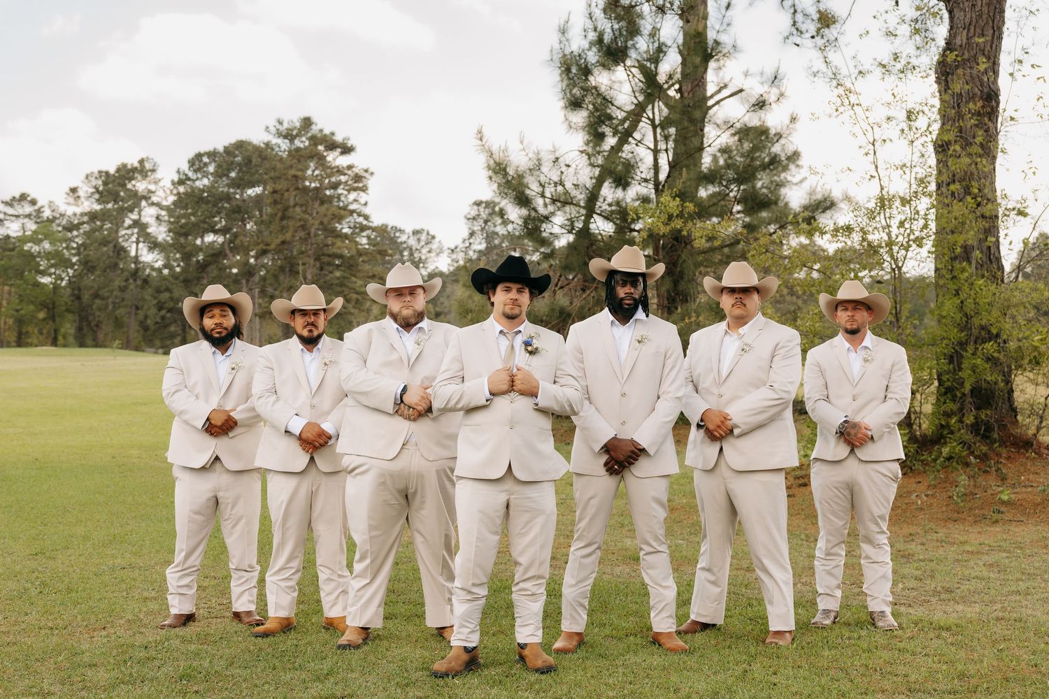 Seven groomsmen in matching cream suits and cowboy hats pose outdoors on a green lawn with pine trees behind them.