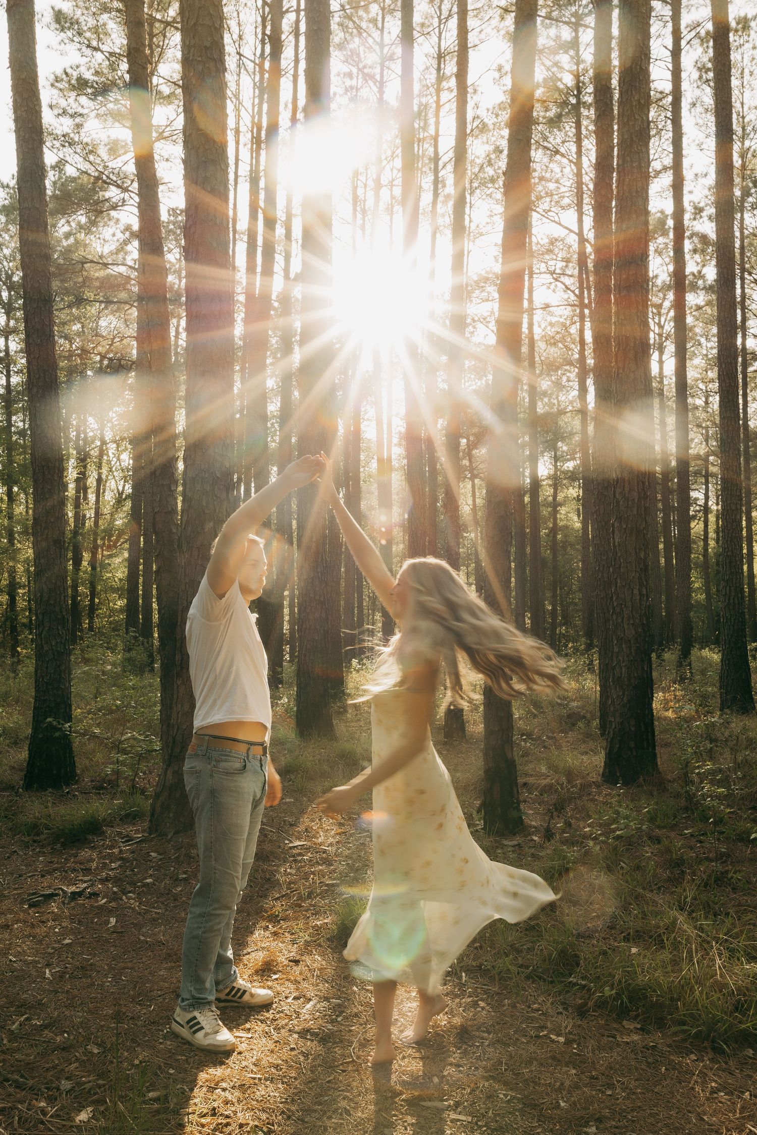 Couple dancing in sunlit forest with dramatic lens flare, woman in flowing dress twirling with partner on woodland path.