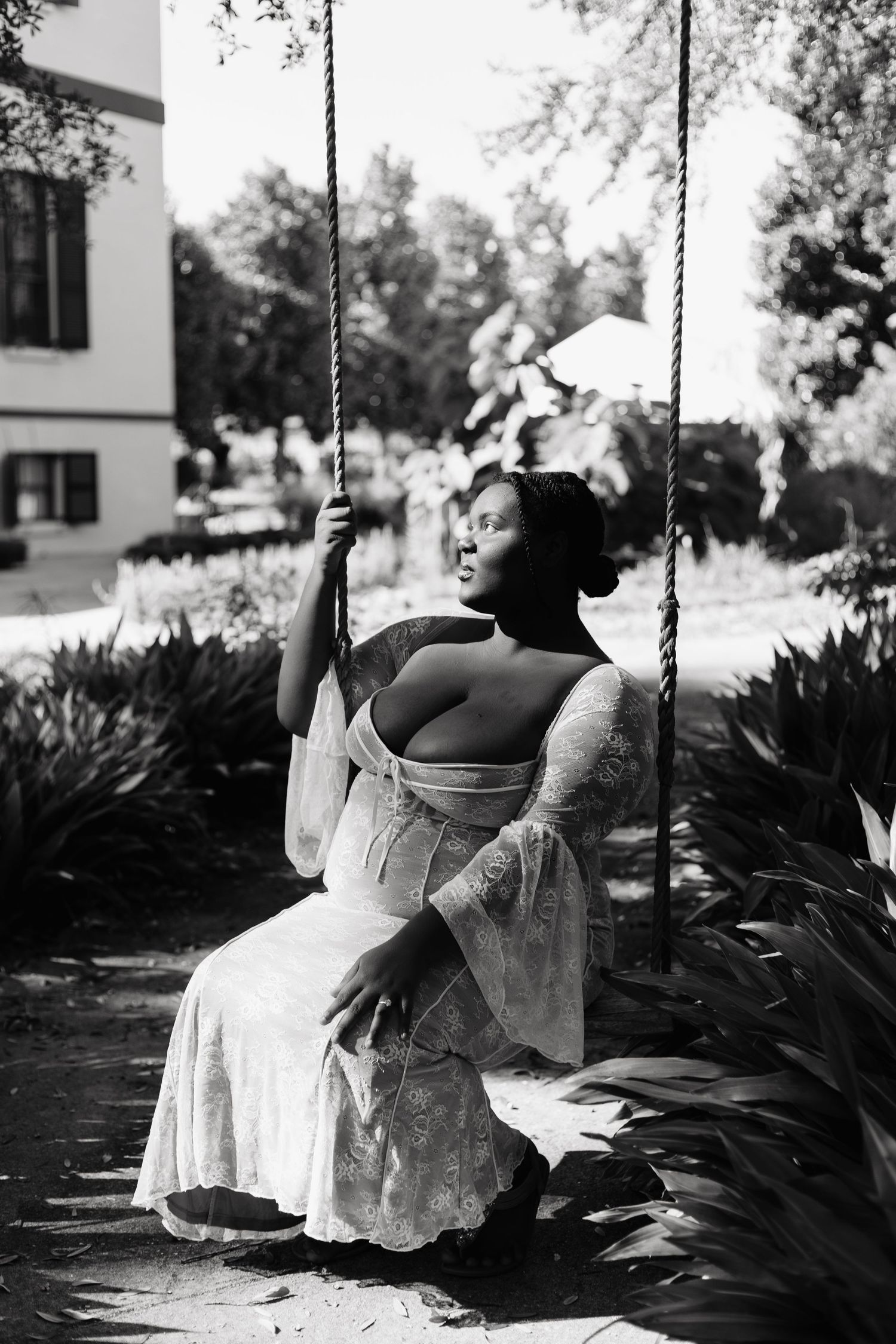 Black and white photo of a woman in a lace off-shoulder dress sitting on a rope swing outdoors.
