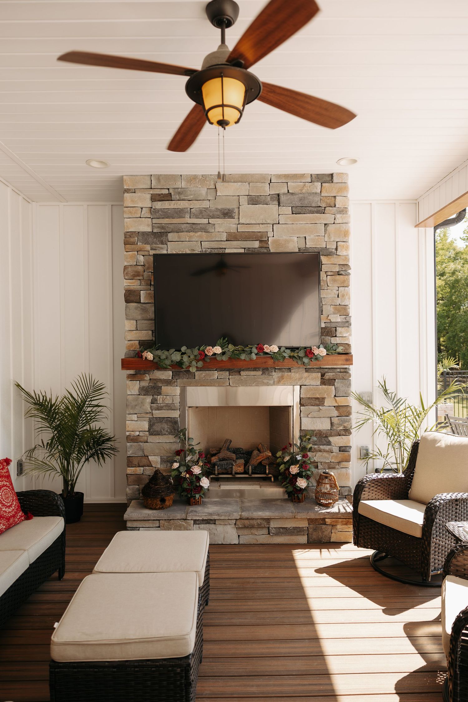 Cozy screened porch with stone fireplace, wall-mounted TV, wicker furniture, and ceiling fan on hardwood floors.