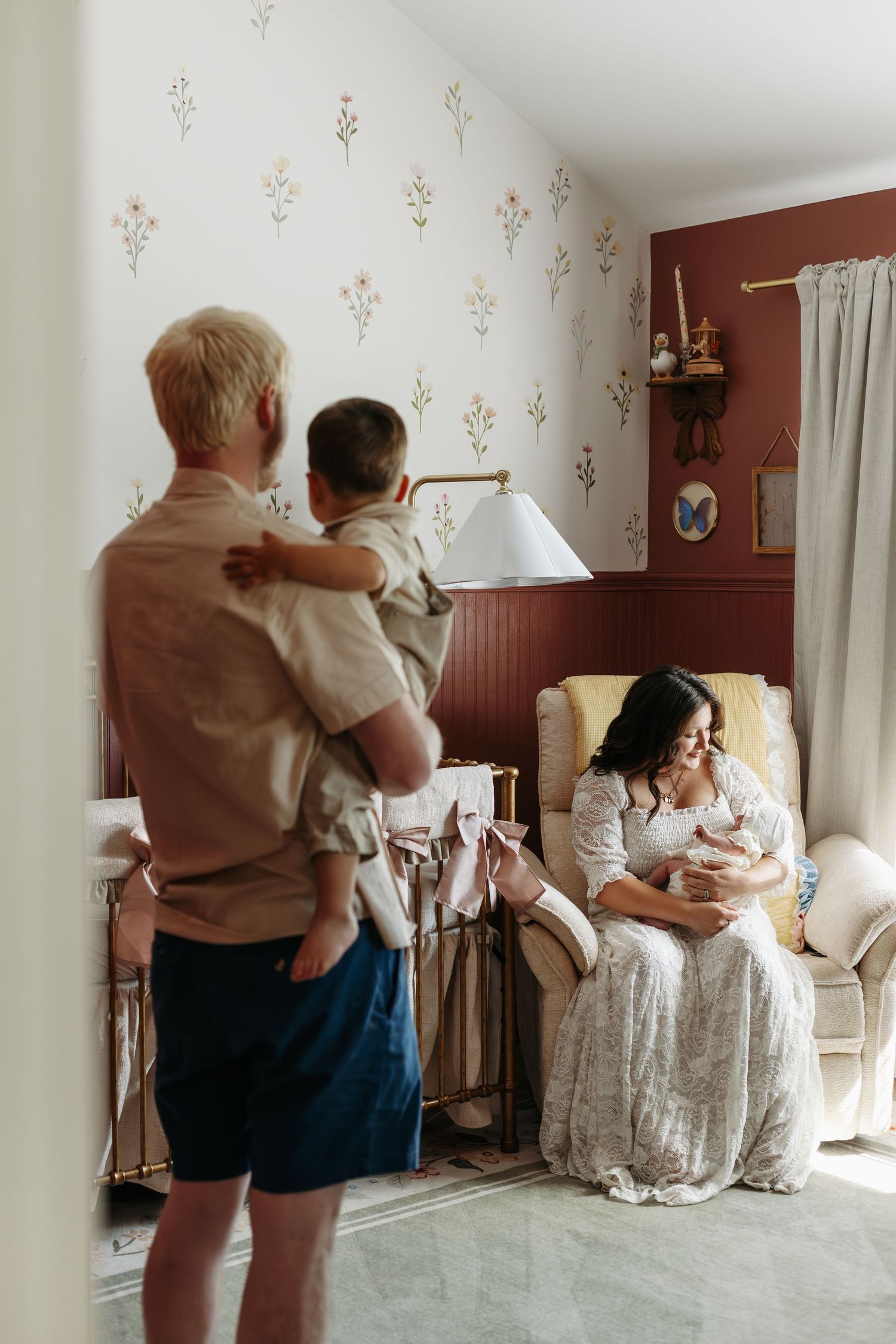A mother in a white dress holds a newborn in a nursery chair while a father holds their toddler nearby.