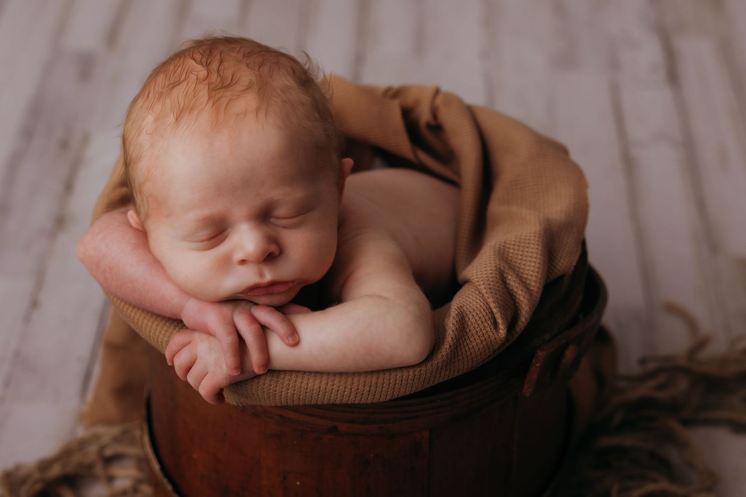 Sleeping newborn baby with red hair posed in a wooden bucket with tan wrap for newborn photography.