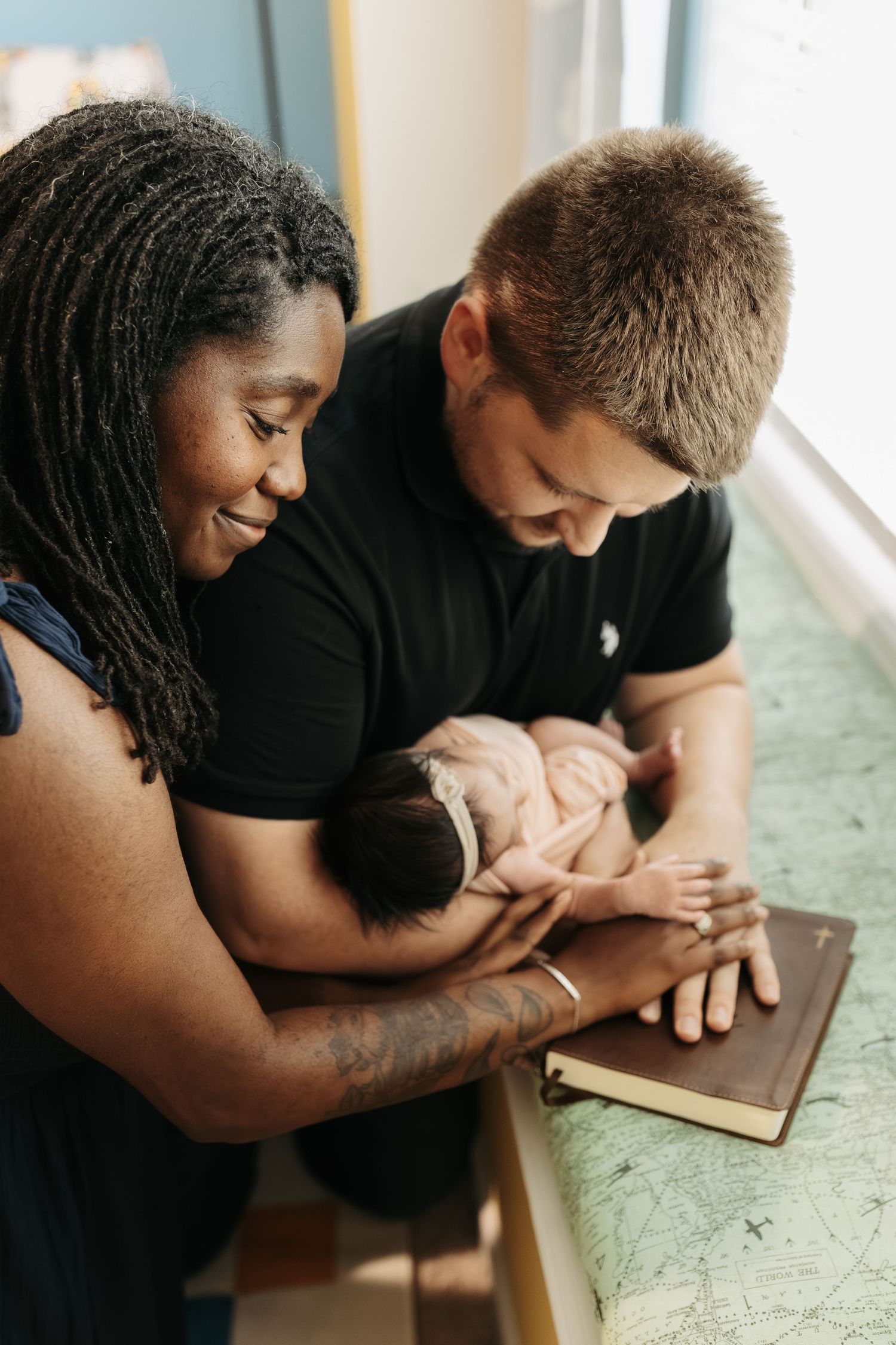 Interracial couple smiling and praying over their newborn baby with a Bible during a church dedication.