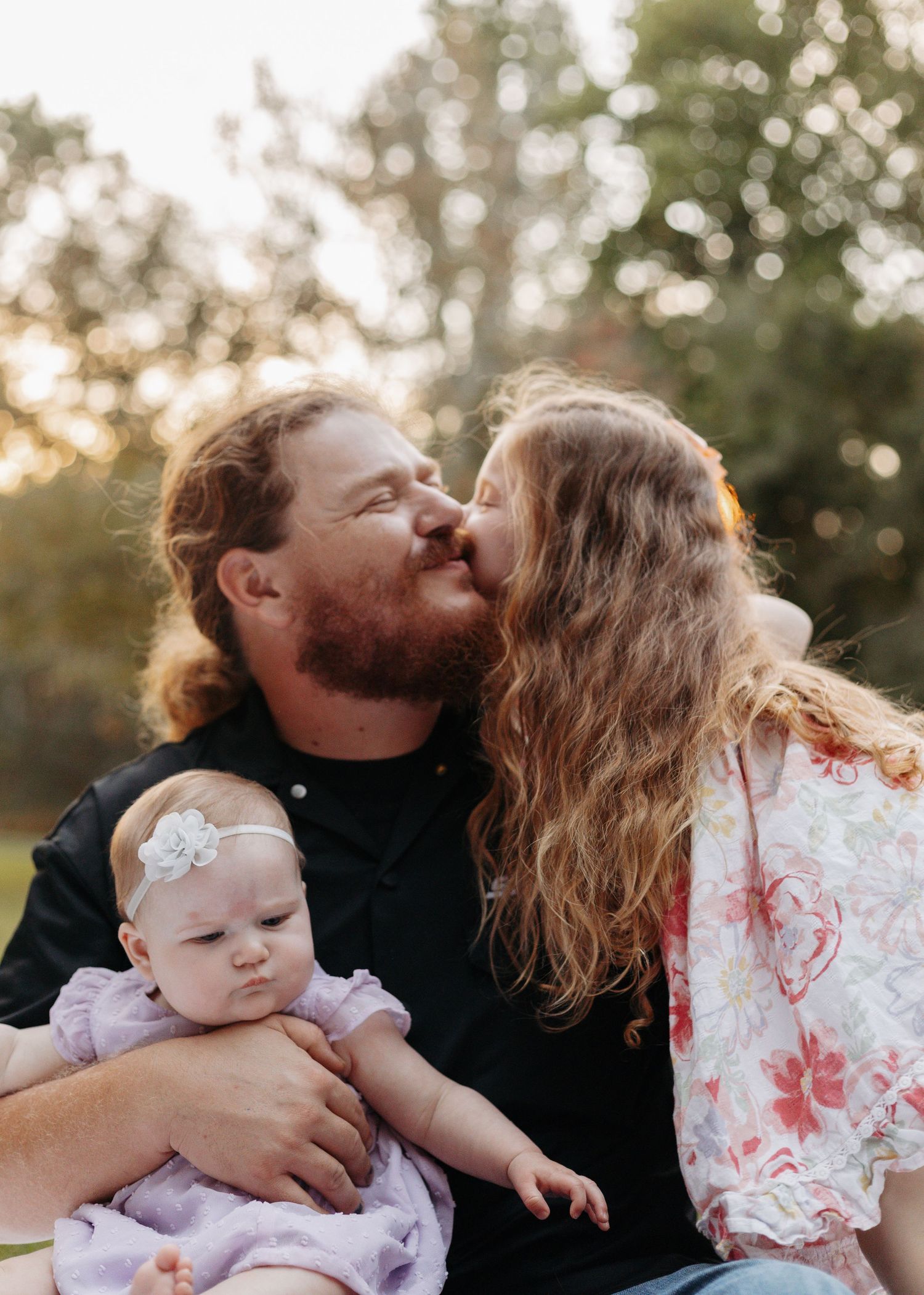 A man holding a baby in purple dress while a girl with long hair kisses his cheek outdoors at golden hour.