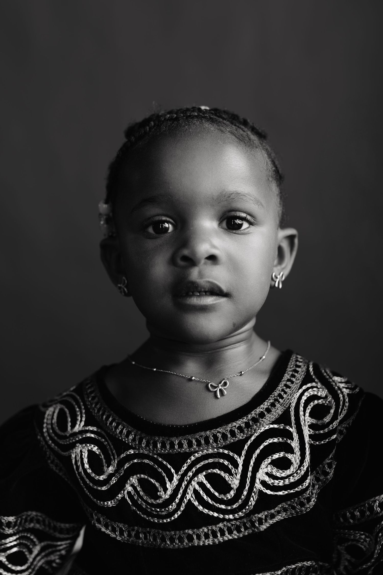 Young African girl in ornate embroidered dress with jewelry, serene expression, black and white studio portrait.