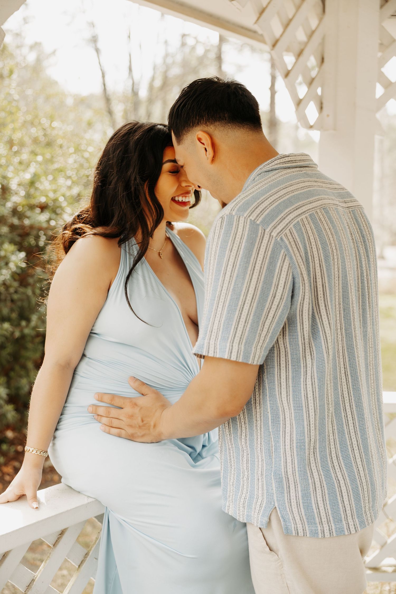 A smiling pregnant woman in a light blue dress shares a tender moment with her partner on a white garden gazebo.