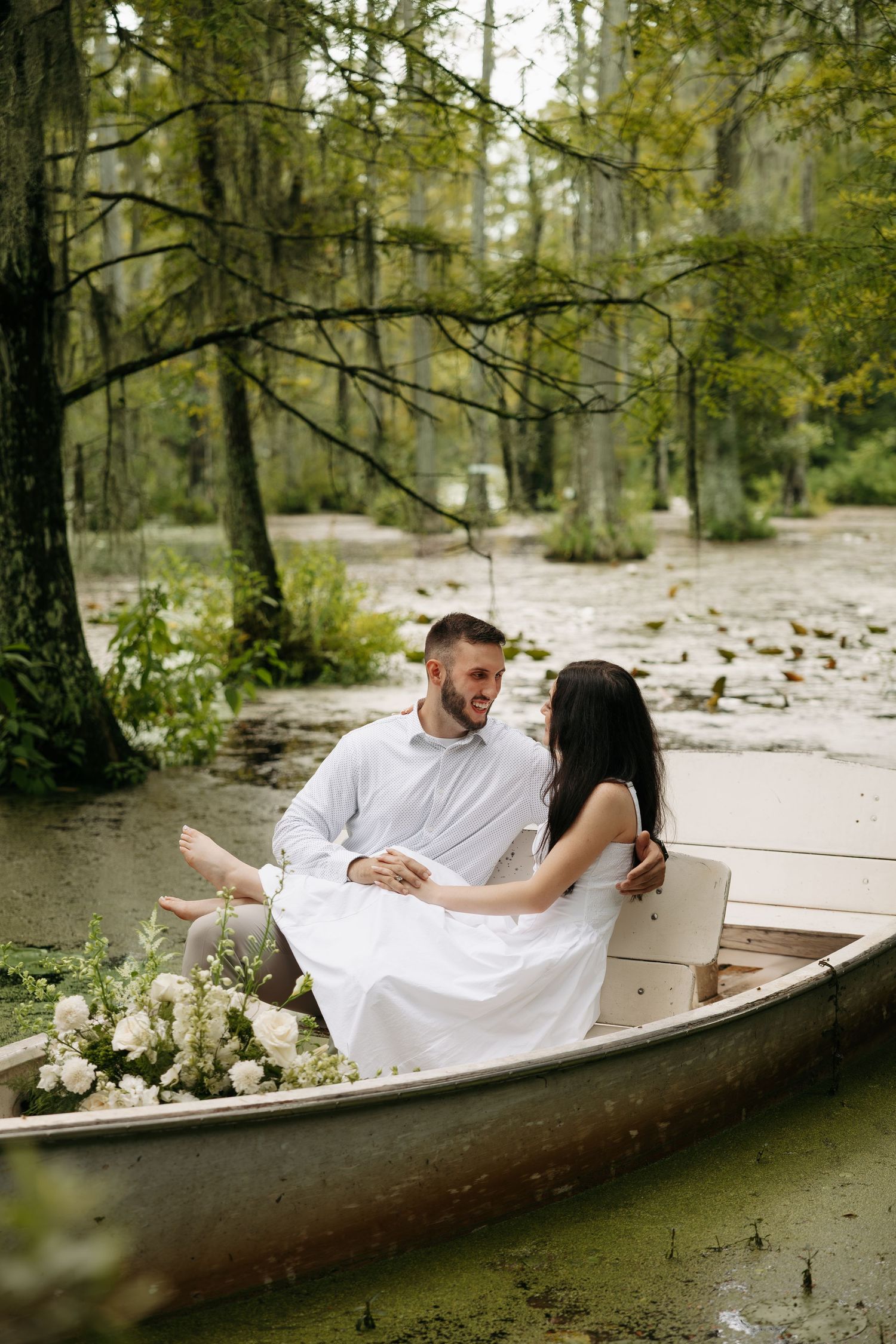 Couple in white outfits sitting romantically in a wooden boat surrounded by lush greenery and white flowers on a serene lake.