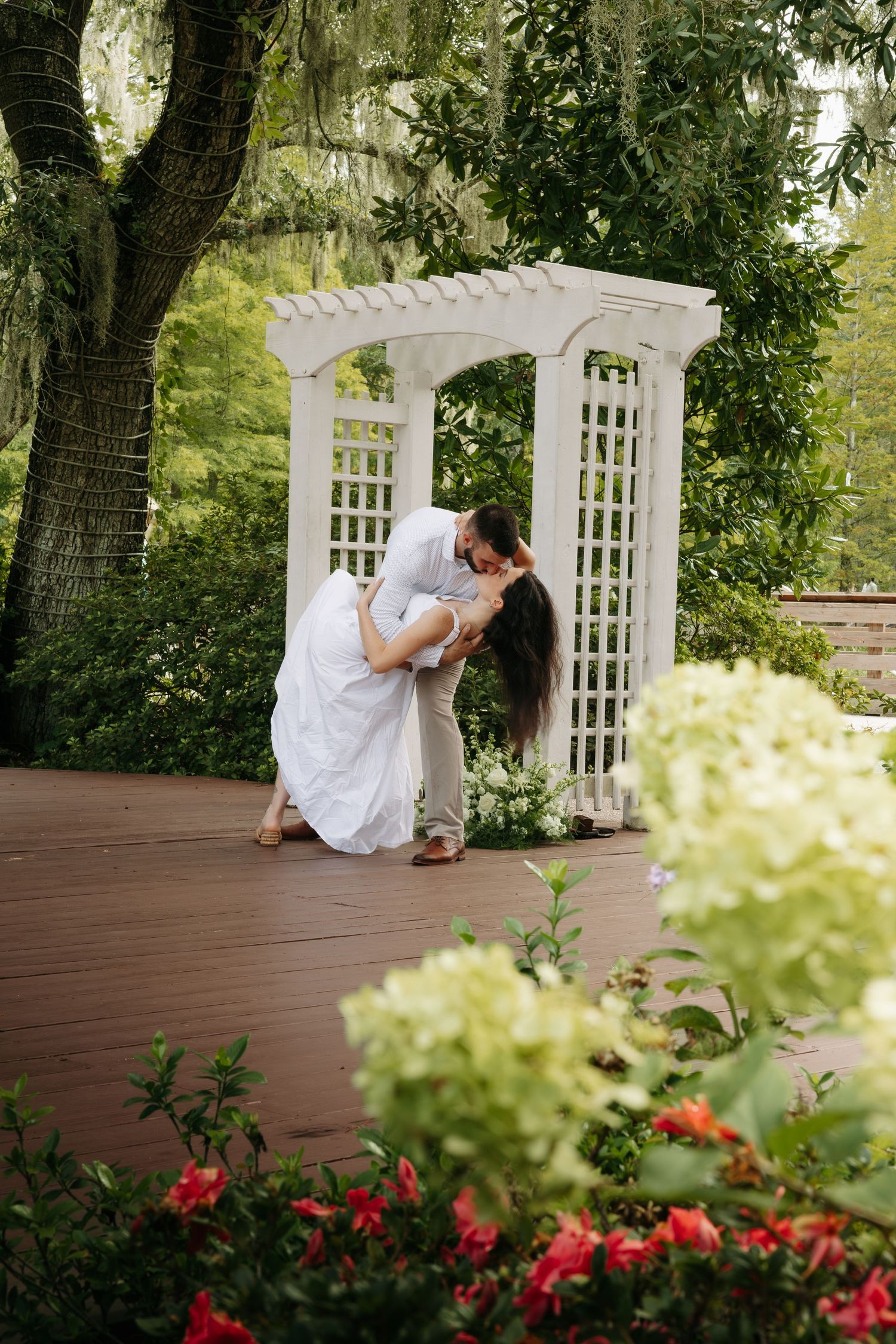 Couple sharing a romantic dip kiss under a white garden arbor on a wooden deck with flowers.