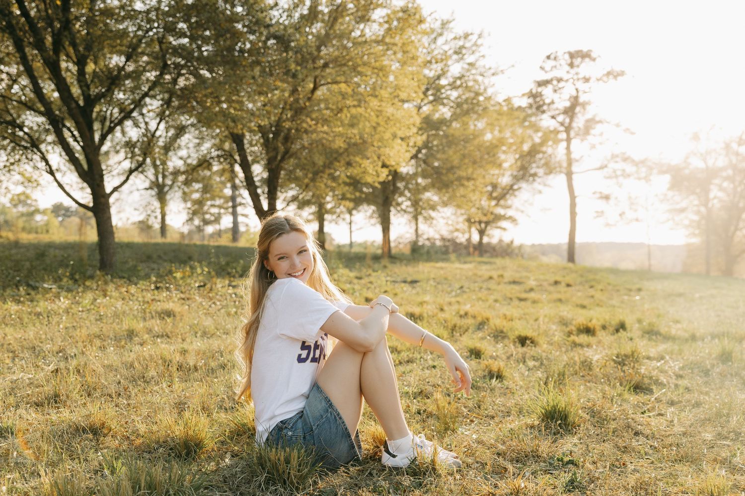 Young woman in white tee and denim shorts sitting on grass in golden hour sunlight with trees in background.