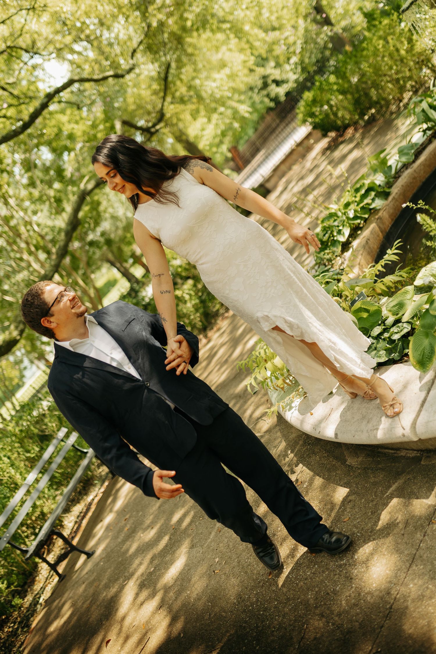 Bride in white gown stands on stone ledge holding groom's hand in navy suit, laughing together in lush green park.