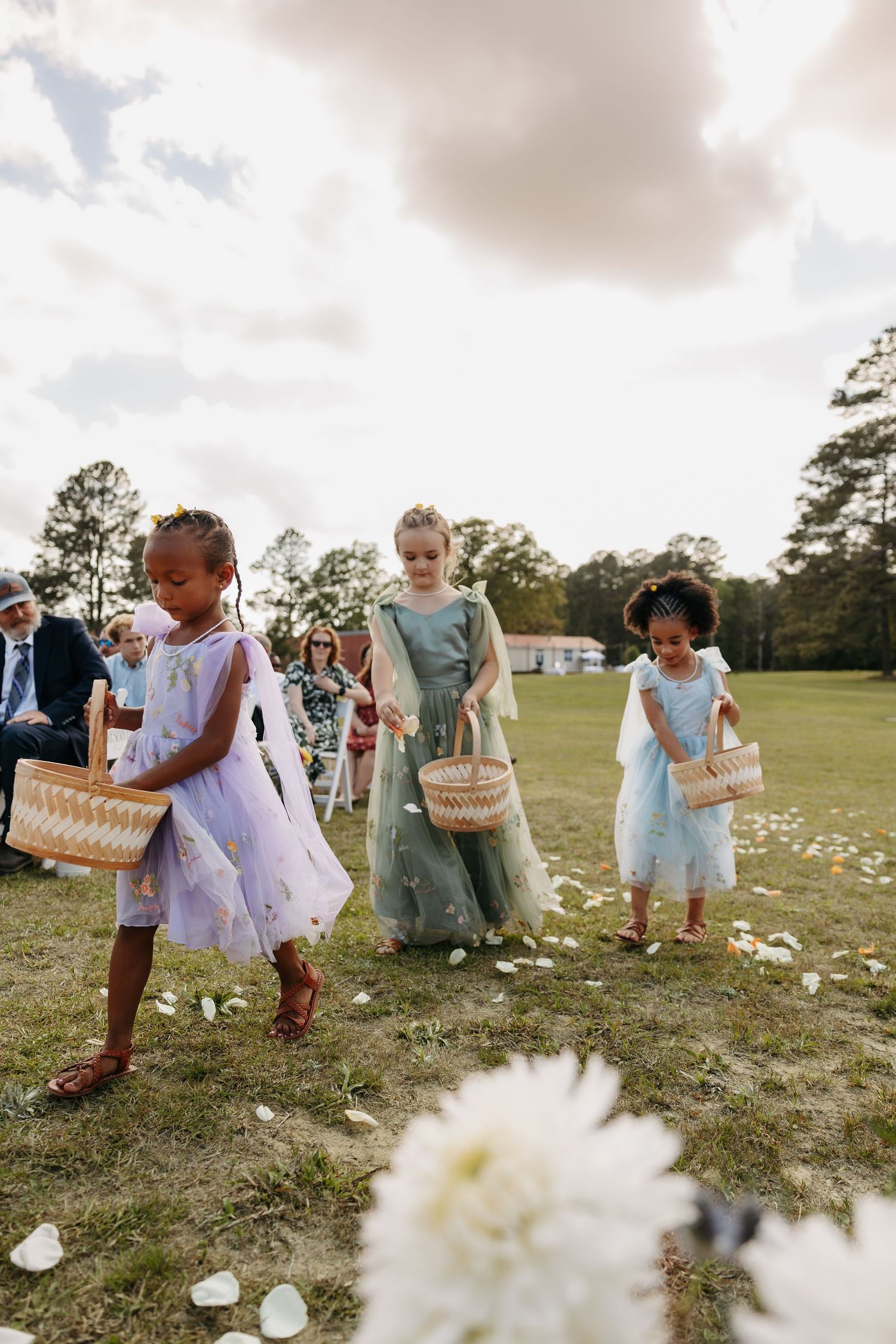Three flower girls in pastel dresses scatter white petals down a grassy wedding aisle outdoors.