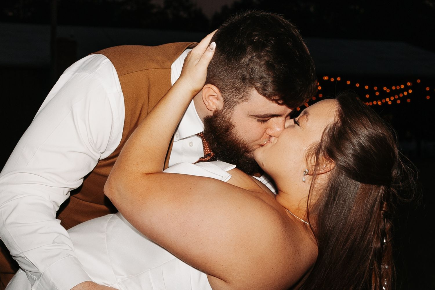 A couple shares a romantic kiss during their wedding reception under warm string lights at night.