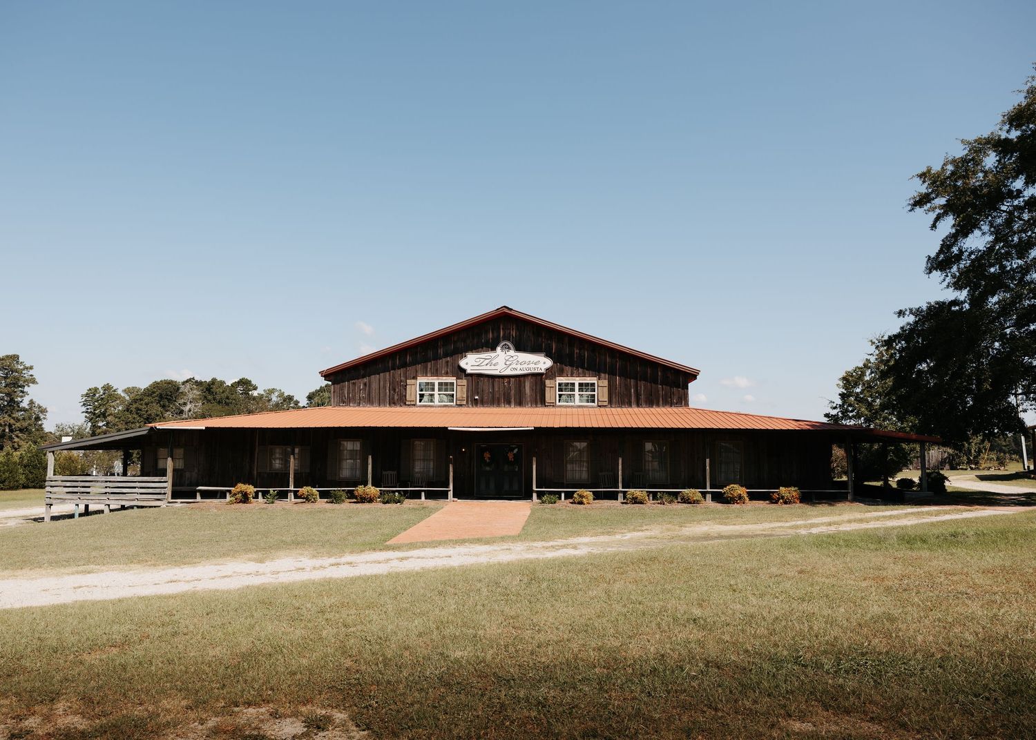 Rustic barn wedding venue with wraparound porch surrounded by green lawns and trees under a clear blue sky.