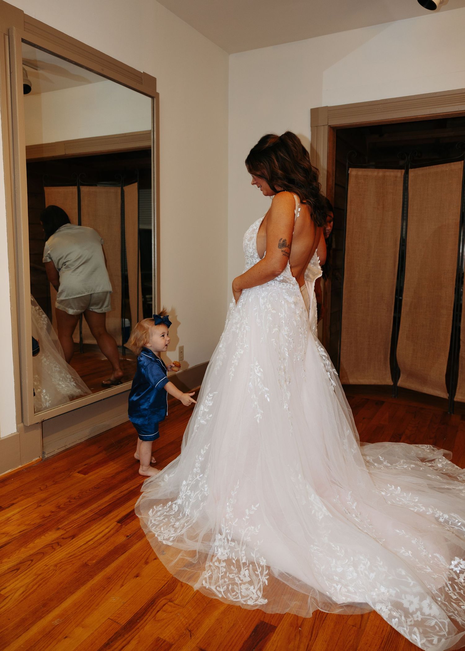 Bride in lace wedding gown stands with toddler flower girl in blue dress before large mirror.