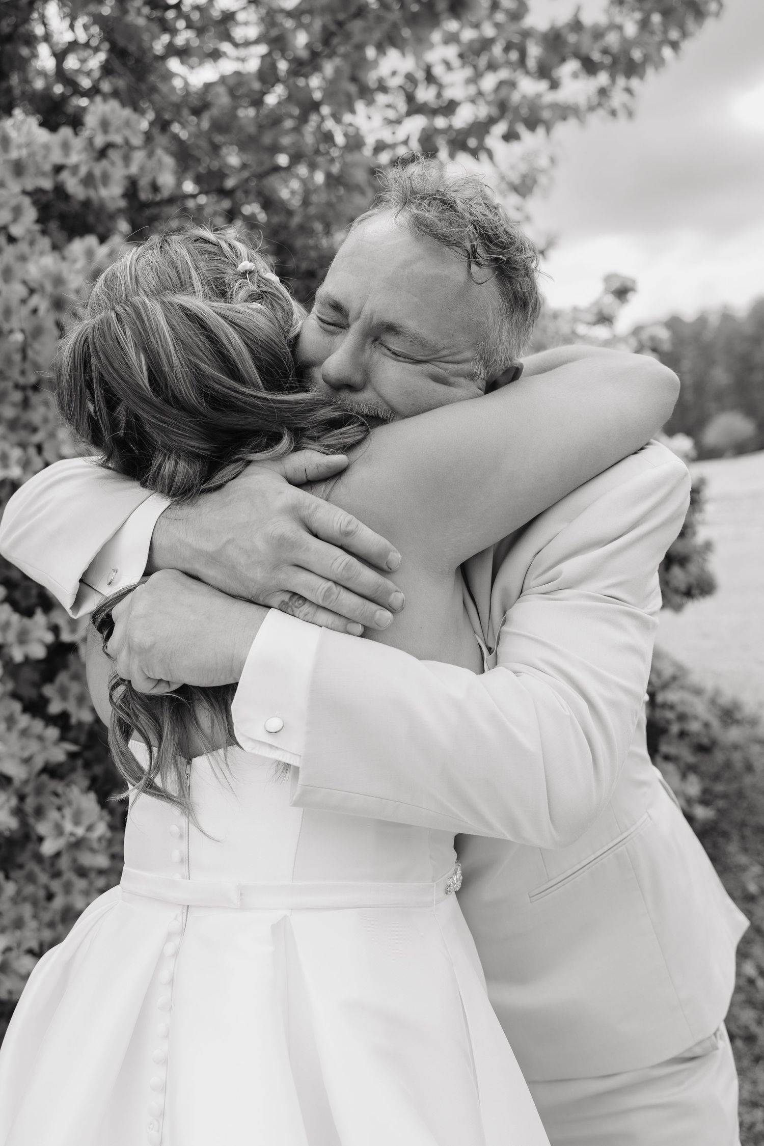 Black and white photo of a bride and older man sharing an emotional hug outdoors at a wedding ceremony.