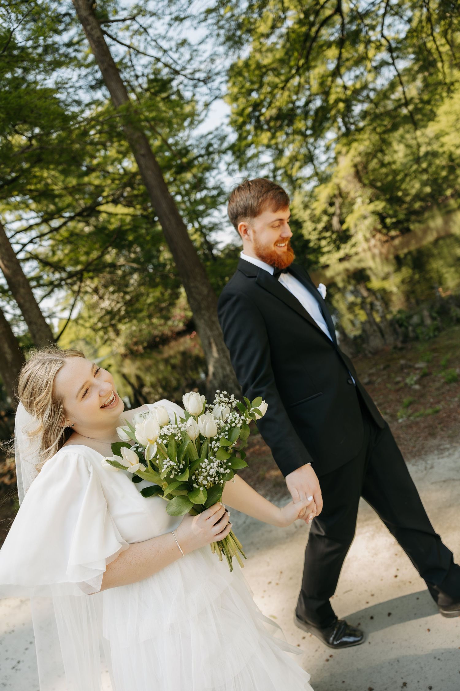 Bride in white dress holding bouquet walks with groom in black tuxedo through a tree-lined path on their wedding day.