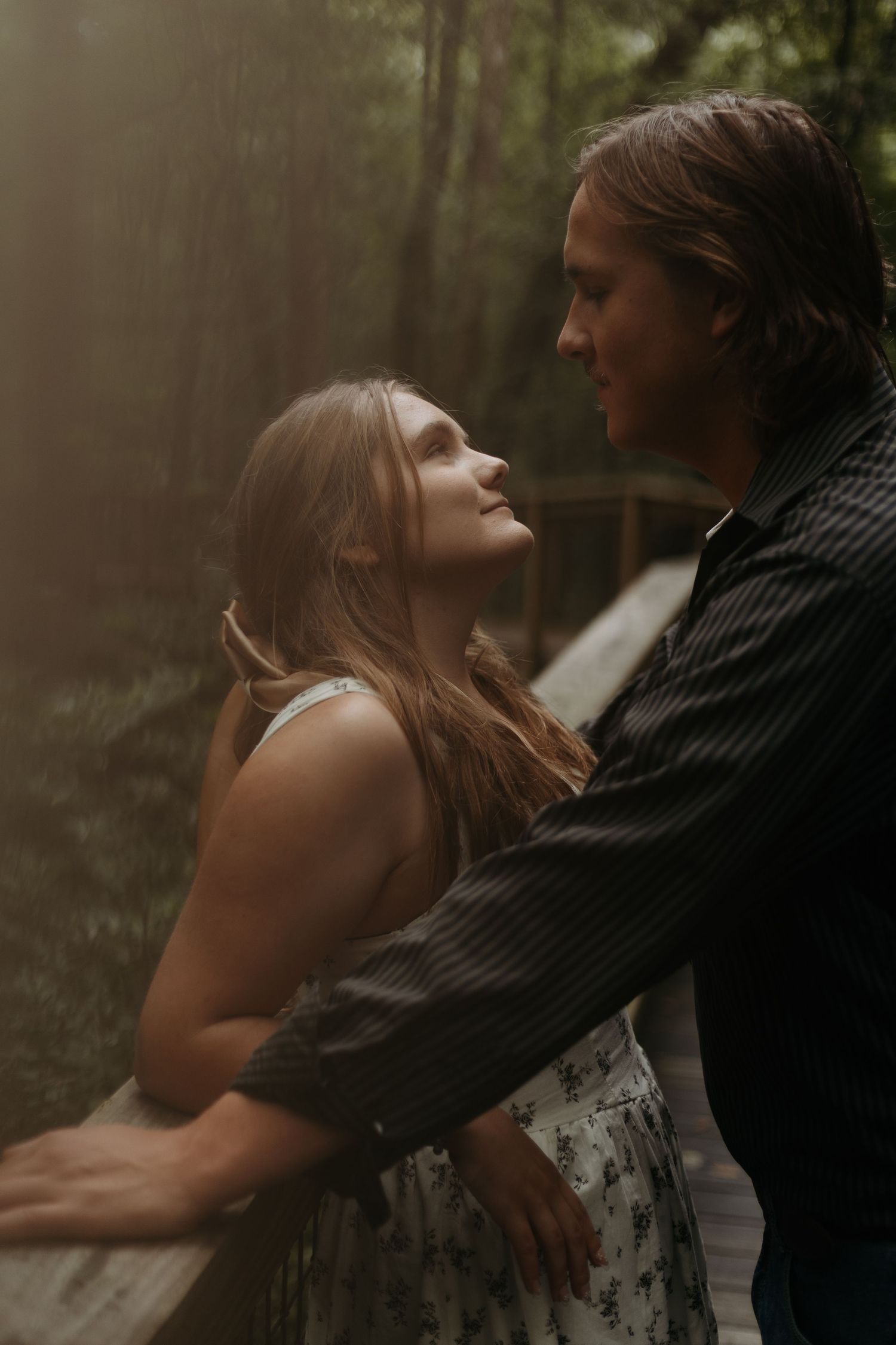 A romantic couple shares a tender moment on a wooden bridge surrounded by misty forest trees in soft golden light.