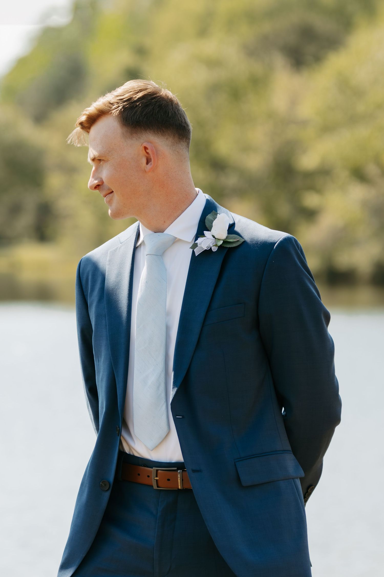 Groom in navy blue suit with boutonniere standing outdoors near a lake on his wedding day.