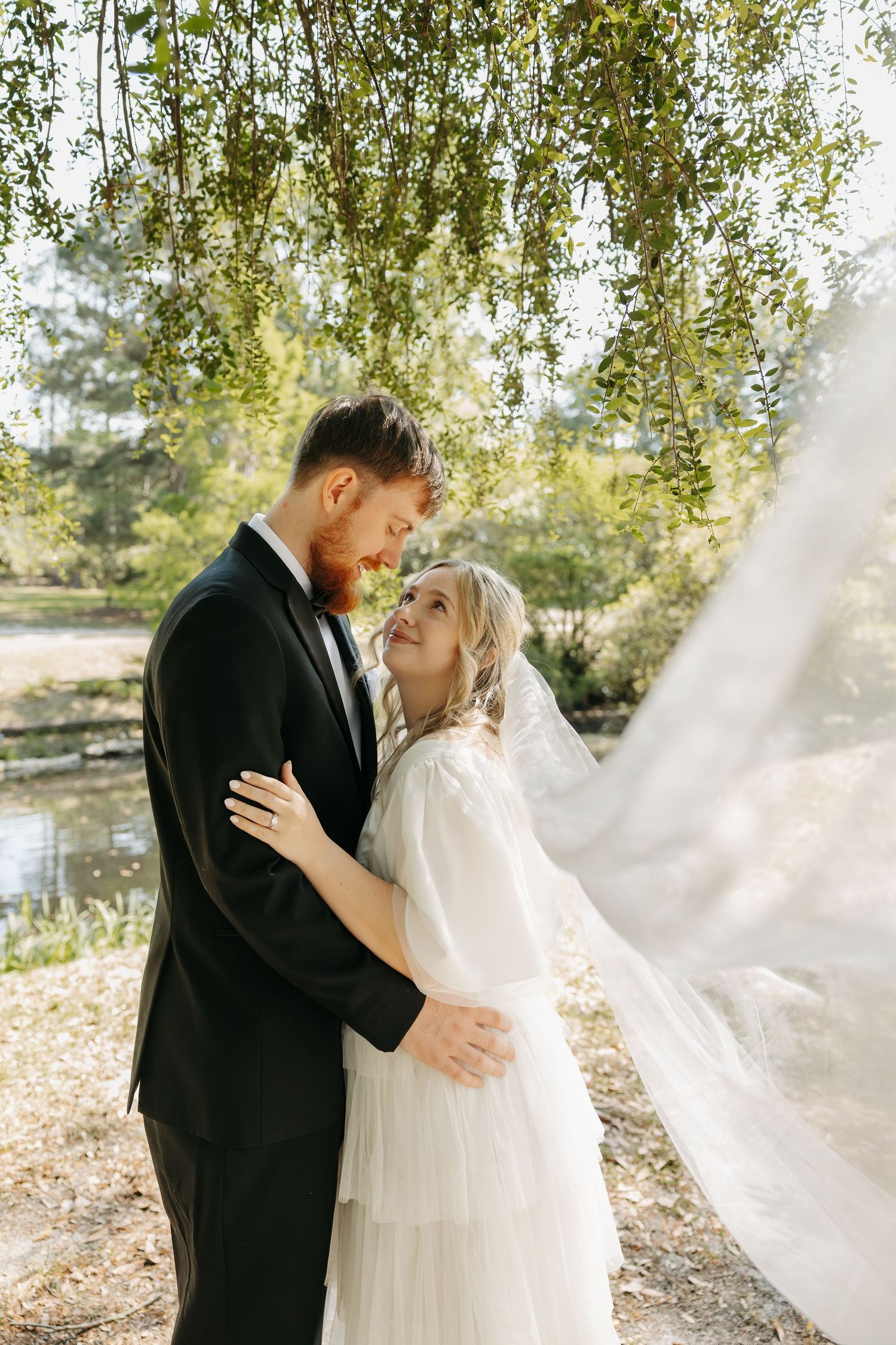 Bride and groom embrace under weeping willow tree by pond, wedding veil flowing in breeze on sunny day.