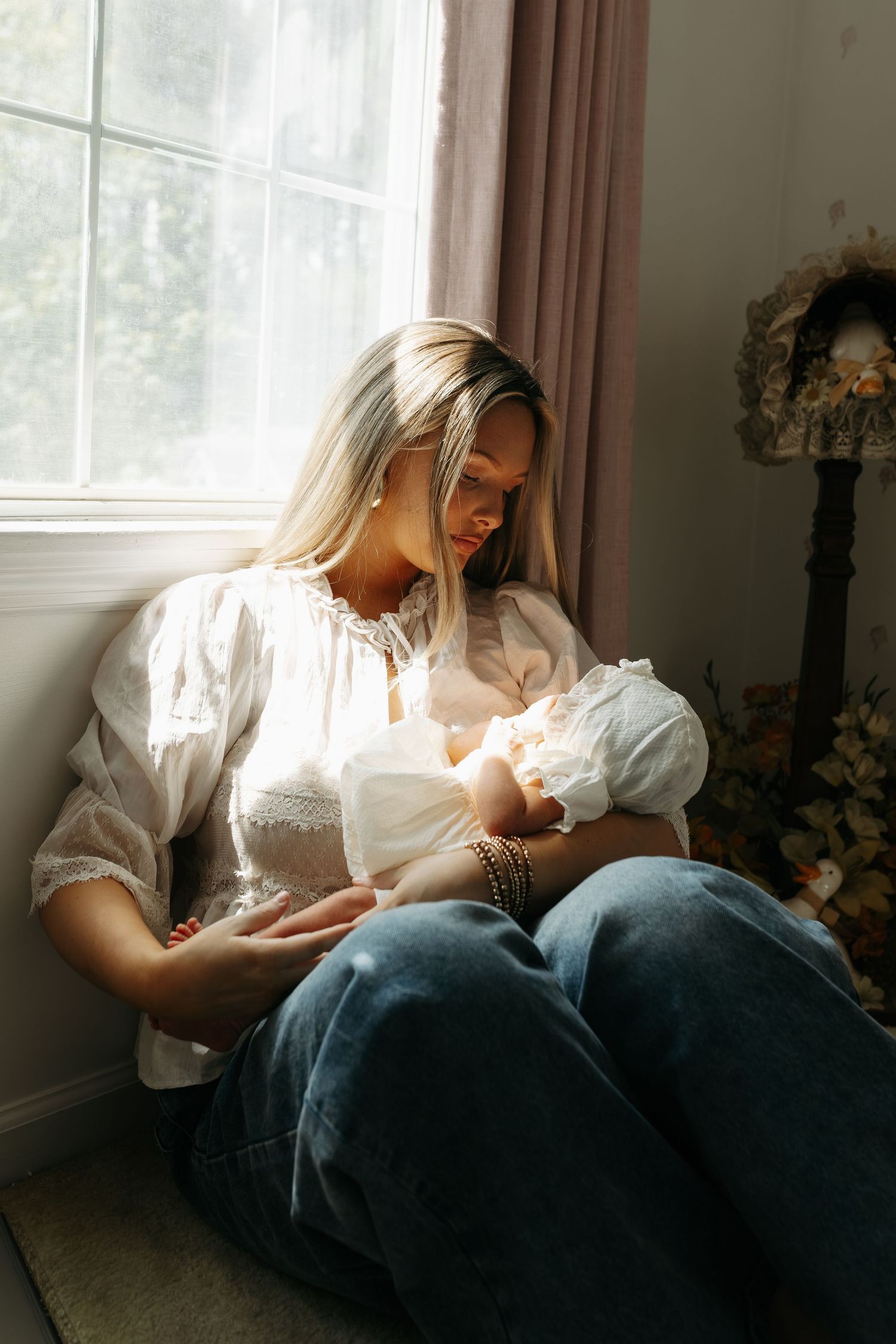 A blonde mother sits by a sunlit window breastfeeding her newborn baby, wearing a white blouse and jeans.