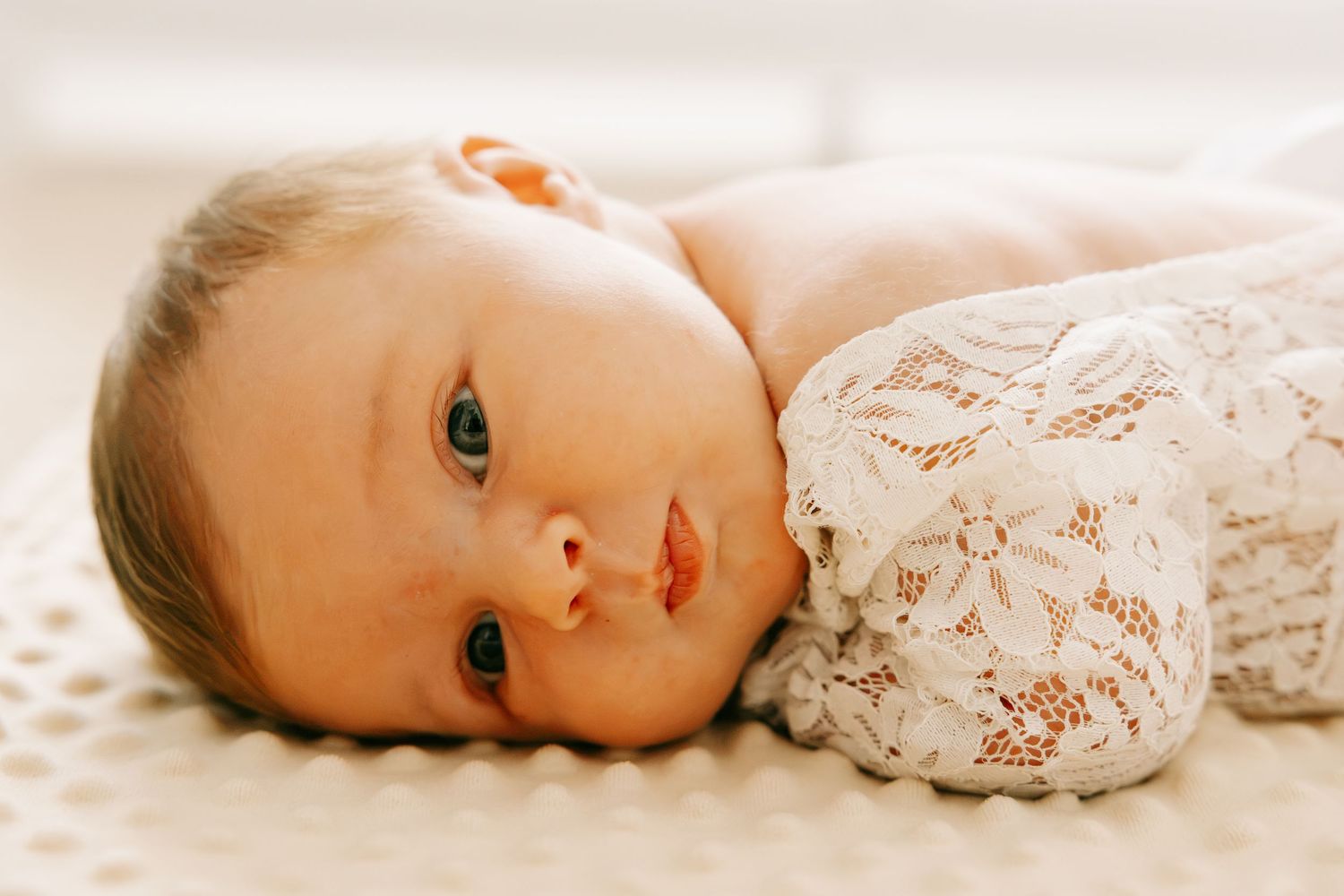 Newborn baby in lace outfit lying on white surface, looking at camera with bright blue eyes.