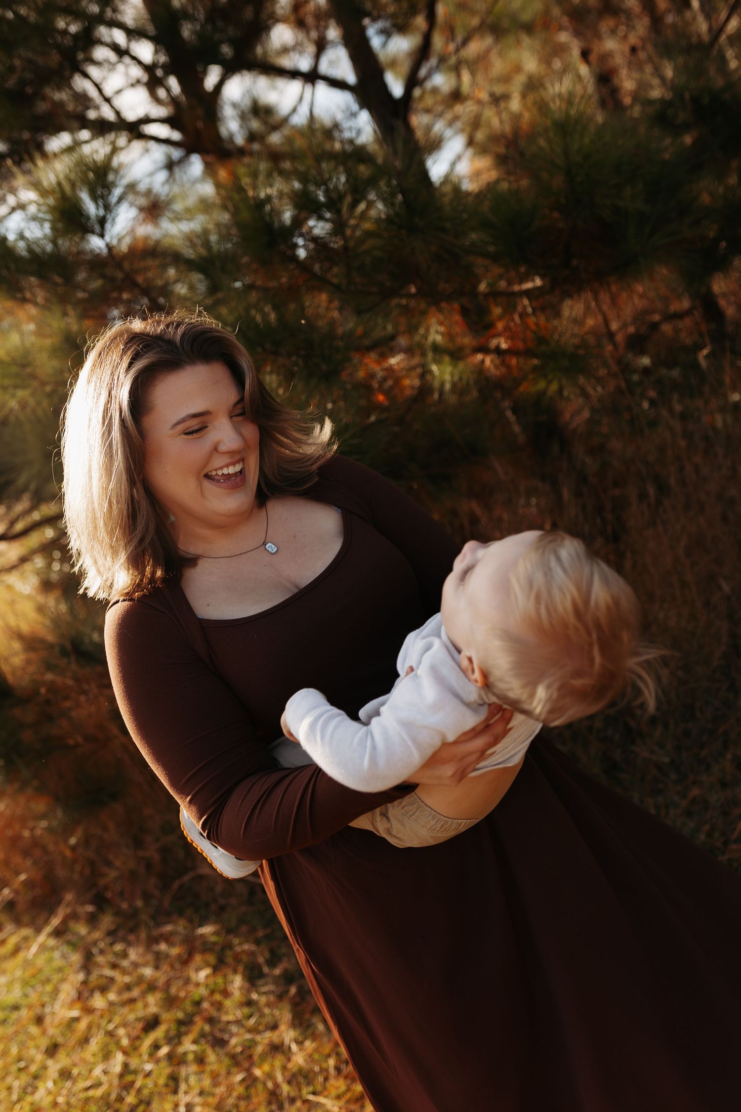 A laughing mother in a brown dress holds her blonde baby outdoors among autumn trees in warm golden light.