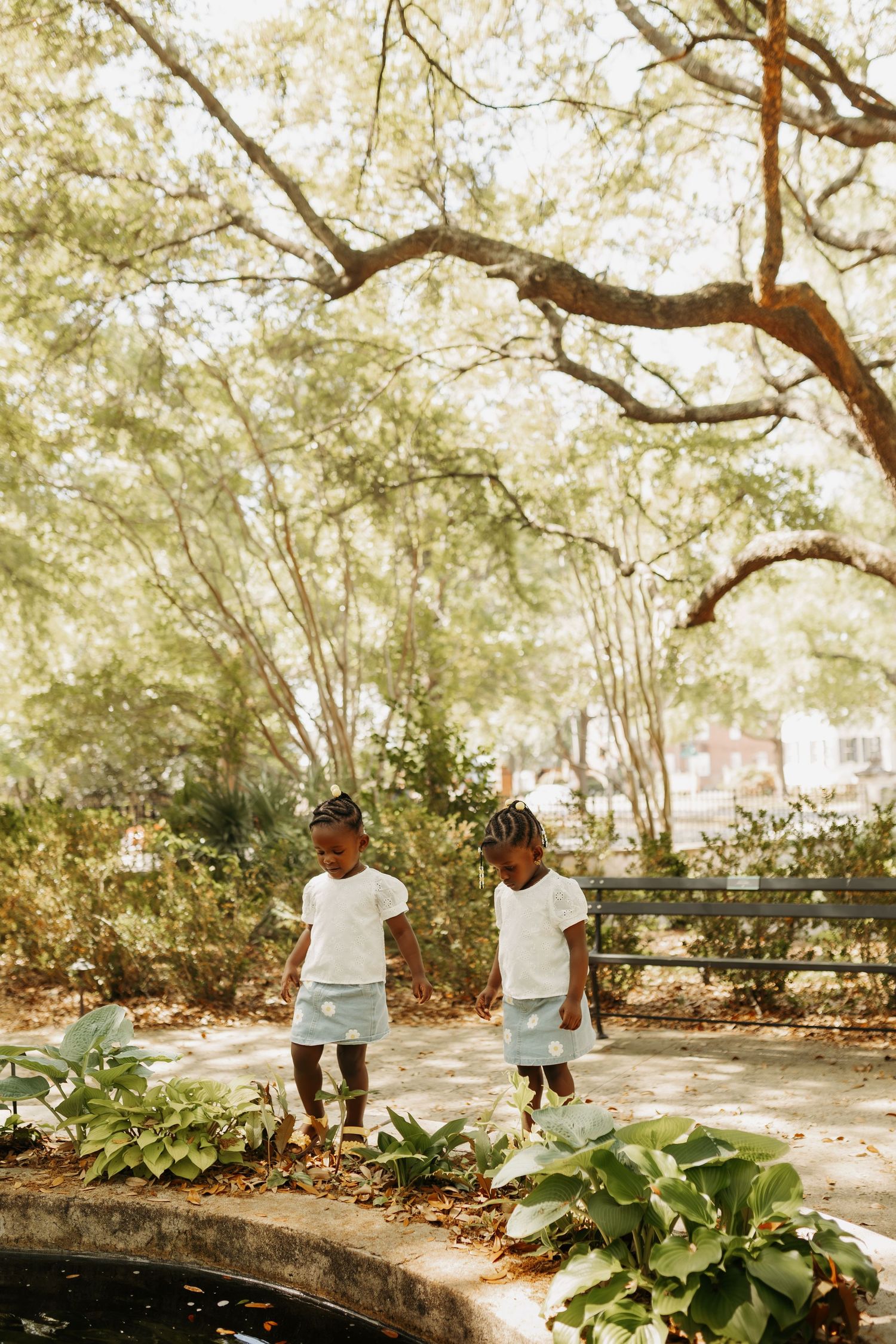 Two young girls in matching white tops and blue skirts stand near a garden pond surrounded by lush green trees.