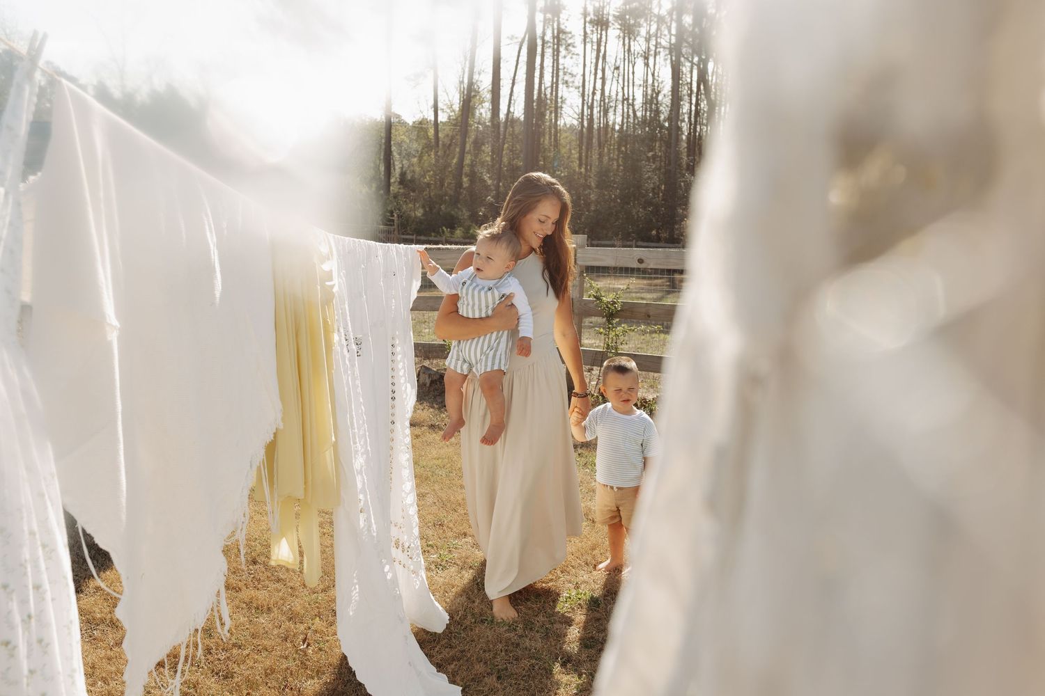 Mother lifting baby outdoors near clothesline with white laundry, toddler standing nearby on sunny farm day.