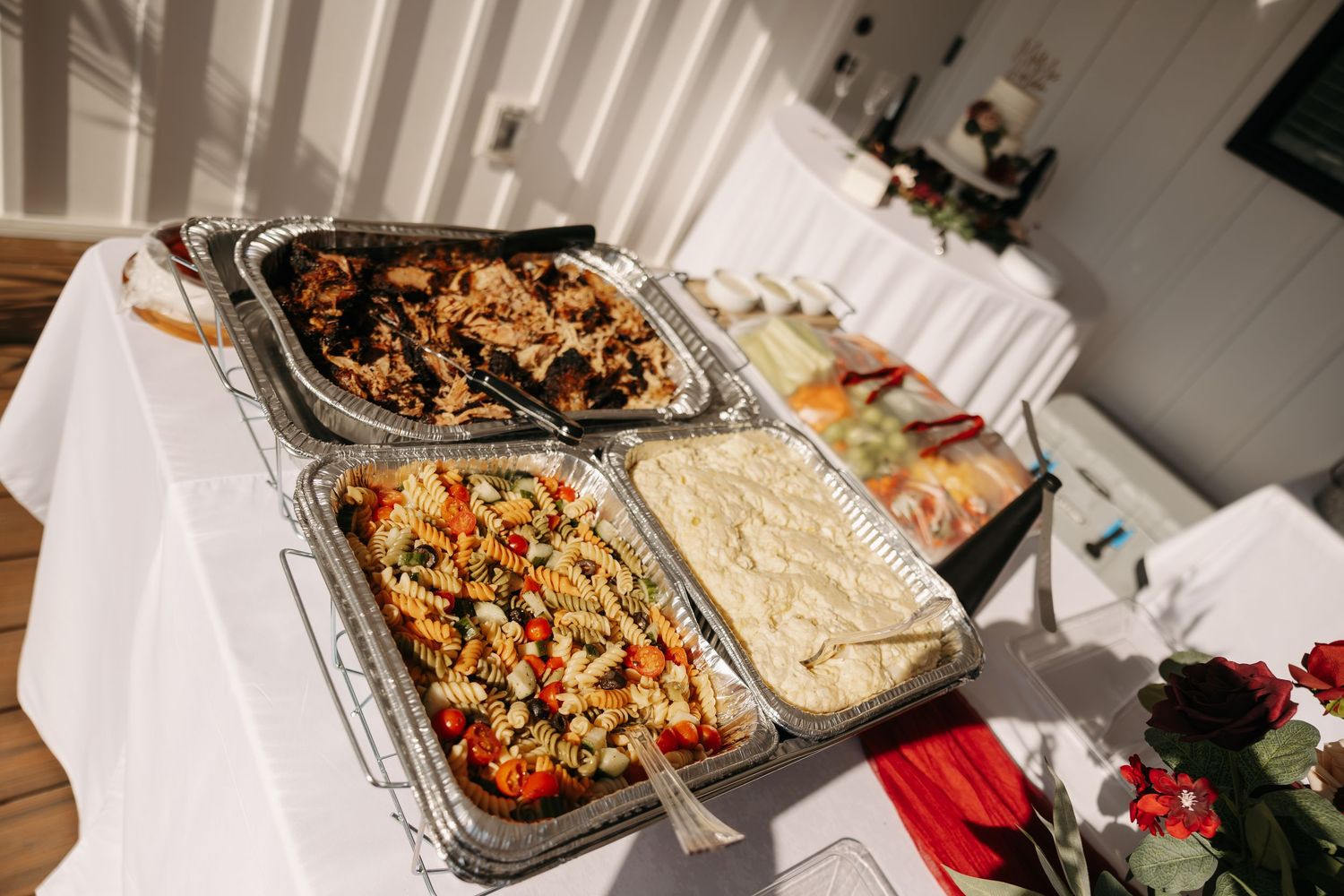 Catered buffet spread with four aluminum trays of pasta, roasted meats, and side dishes on a white tablecloth.