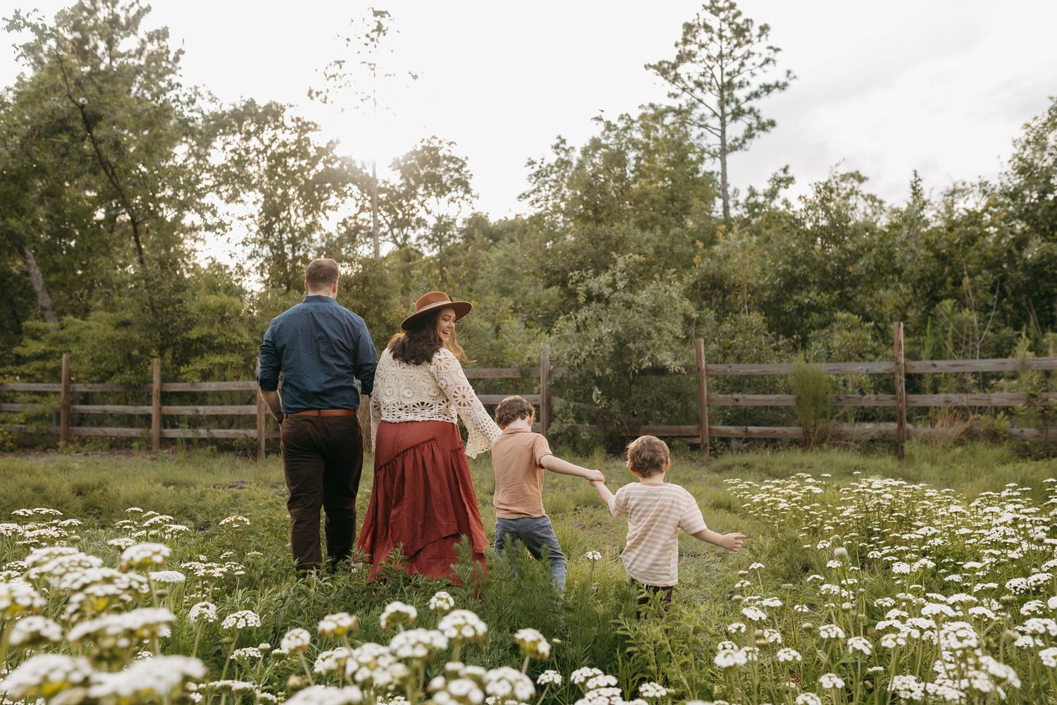Family of four walking through a wildflower meadow holding hands, with a wooden fence and lush trees in the background.
