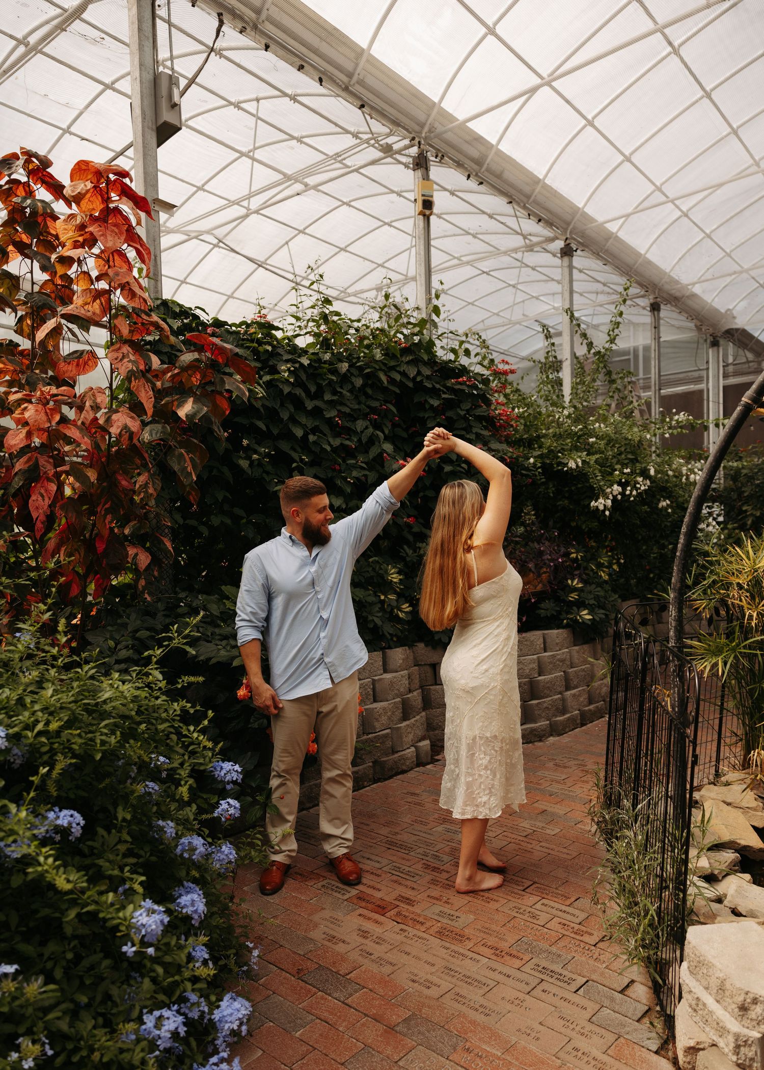 Couple dancing together in a lush greenhouse with brick path, colorful foliage, and blue flowers.