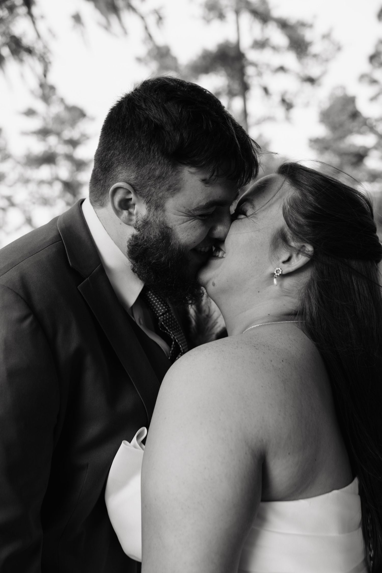 Black and white photo of a bearded groom in a suit kissing his smiling bride in a white dress outdoors.