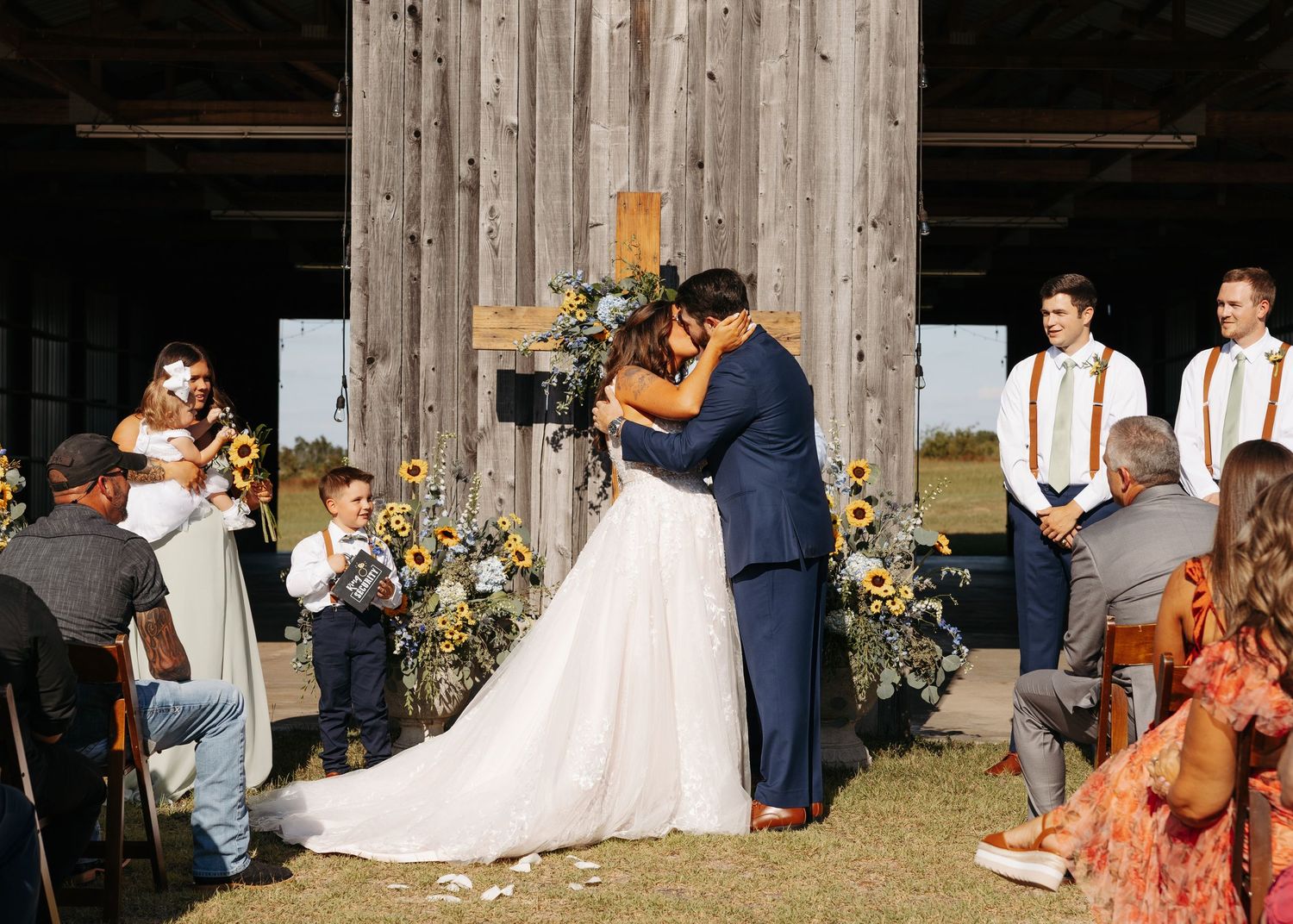 Bride and groom share first kiss at rustic barn wedding altar adorned with sunflowers.