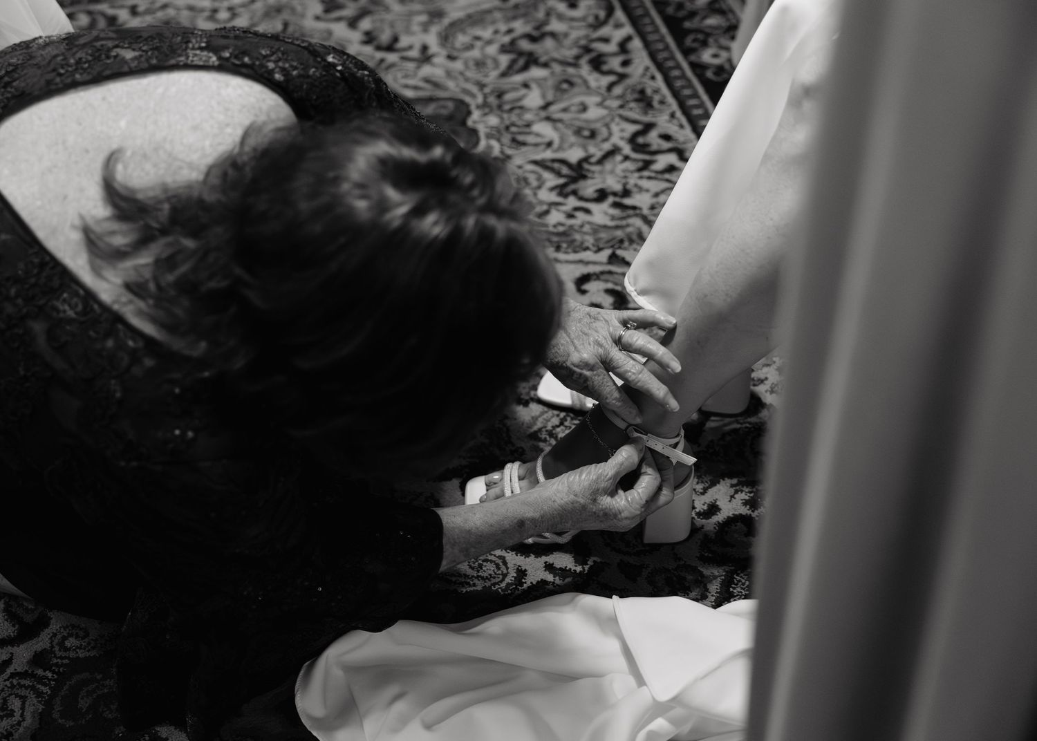 Black and white photo of a woman kneeling to fasten a bridal shoe on a wedding dress hem.