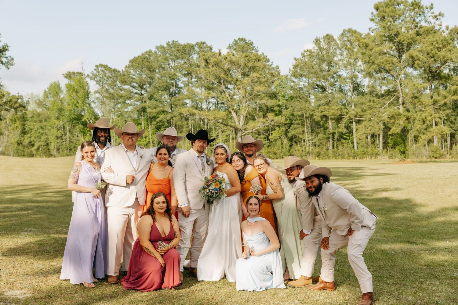Wedding party group photo on a green lawn with bridesmaids in colorful dresses and groomsmen in cream suits.