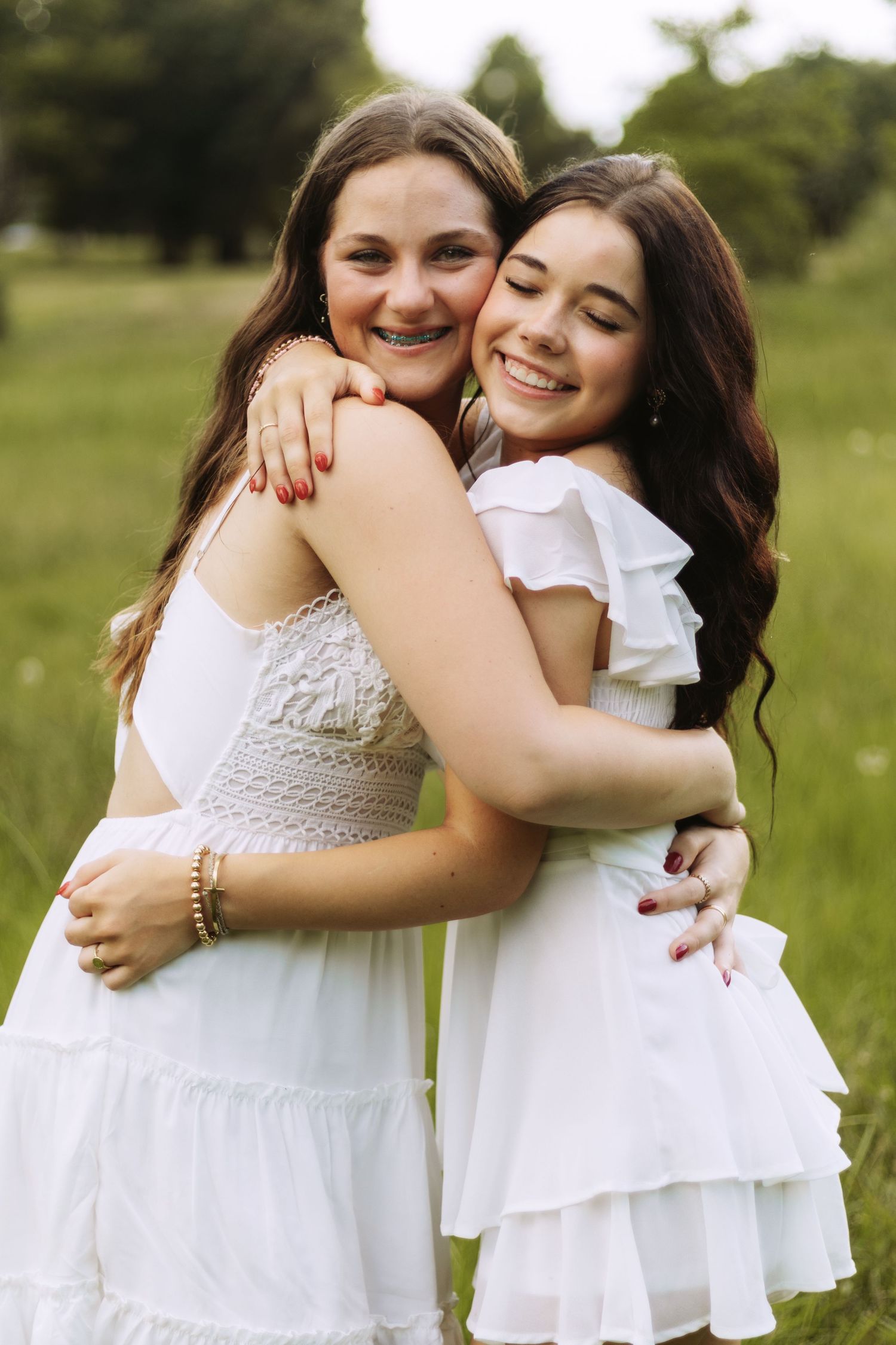 Two people in white dresses embrace and smile together in a sunny outdoor field with greenery in the background.