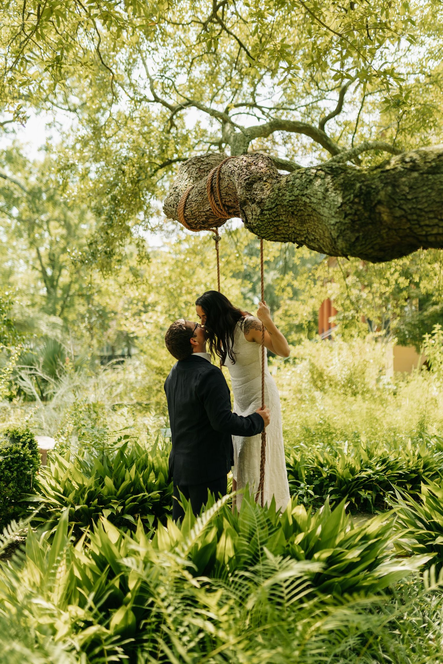 Bride and groom share a romantic moment under a large tree surrounded by lush green ferns in a garden.