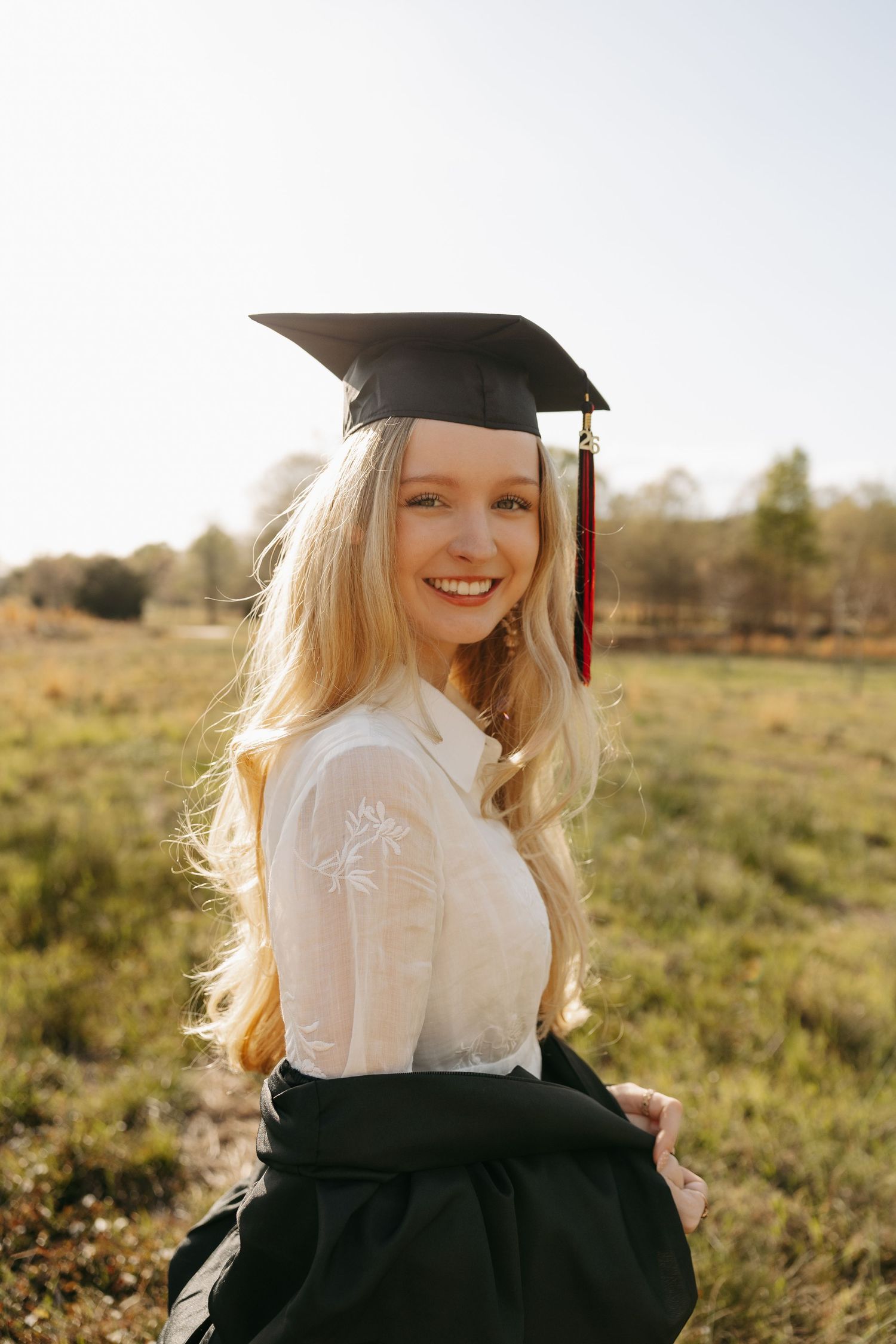 Smiling blonde graduate wearing black mortarboard cap and white blouse in sunny outdoor field setting.