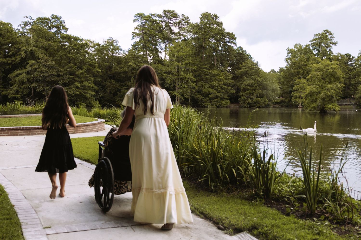 Two people walk along a paved path beside a scenic lake with cypress trees and cattails on a summer day.