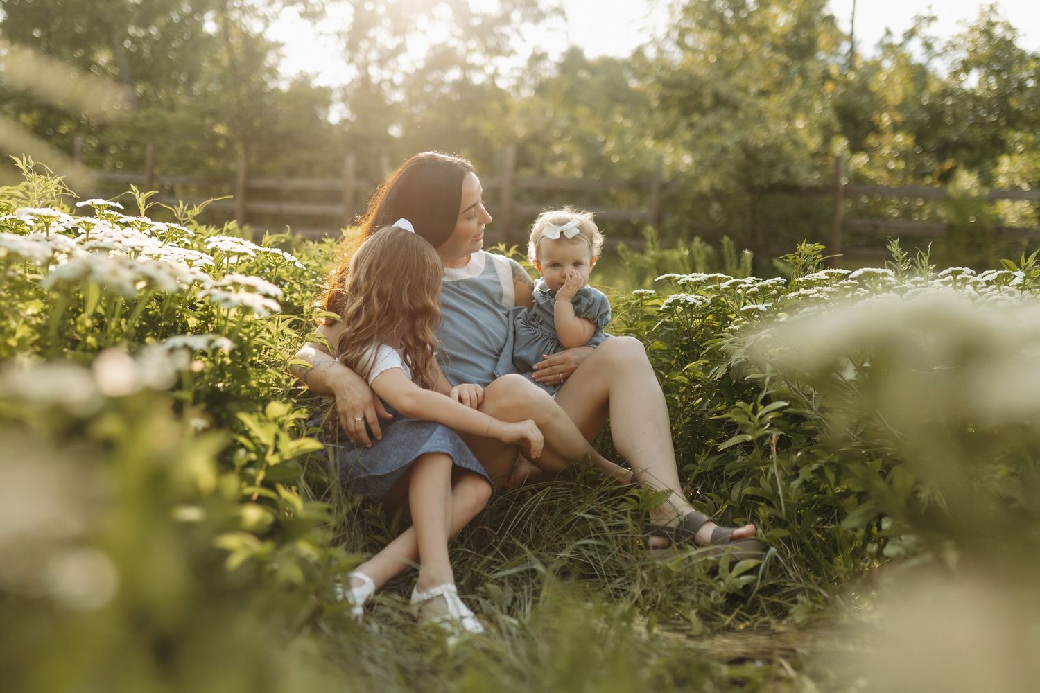 A mother sits outdoors in a sunny garden with her two young daughters surrounded by green plants and white wildflowers.