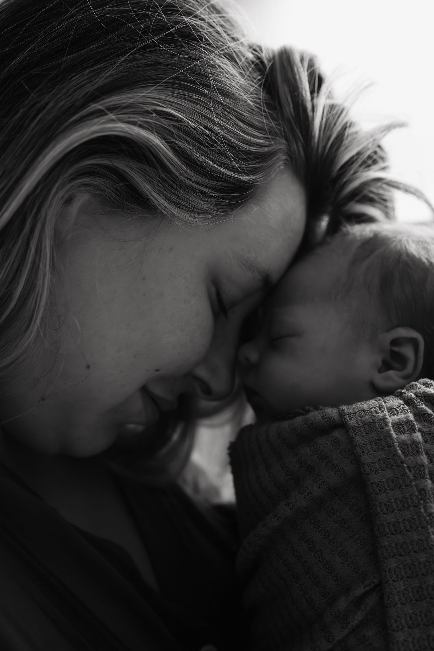 Black and white photo of a mother tenderly touching foreheads with her sleeping newborn baby.