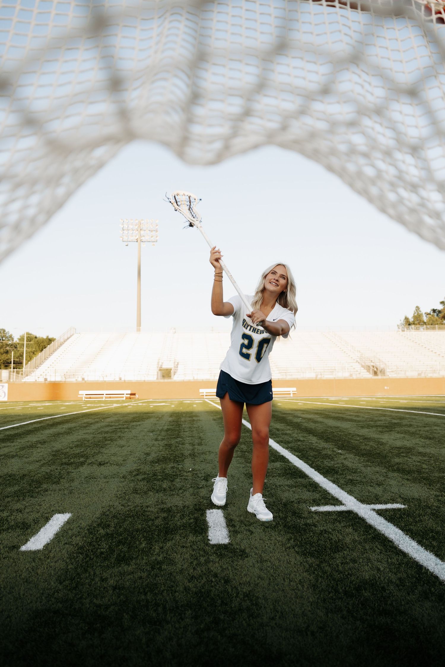 Female lacrosse player in white jersey number 20 holding stick on green field, framed by goal net.
